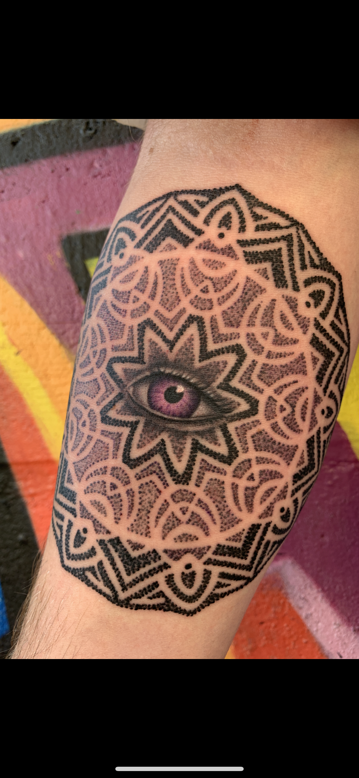 Tattoo of an eye with a purple iris surrounded by intricate black geometric patterns on a person's arm.