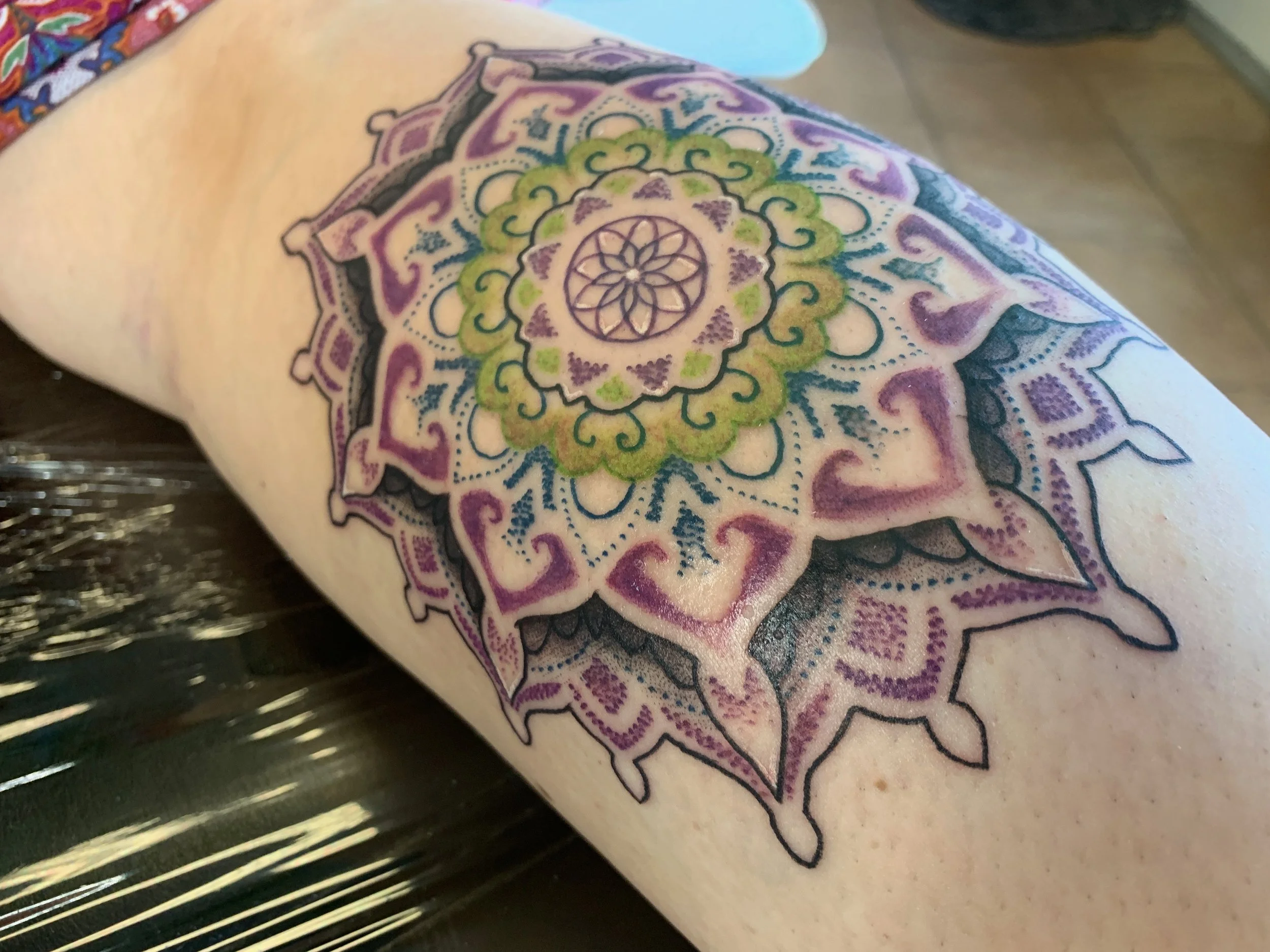 Colorful mandala tattoo on a person's arm featuring intricate patterns in purple, green, blue, and black ink.