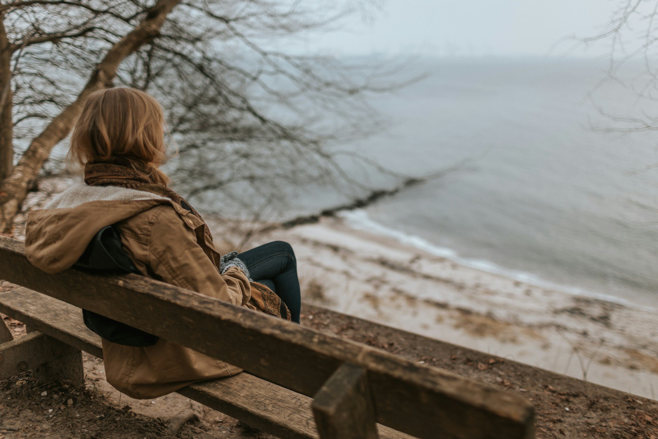 A person with reddish hair wearing a brown jacket and scarf sitting on a wooden bench by the lake, looking at the water with bare trees around.
