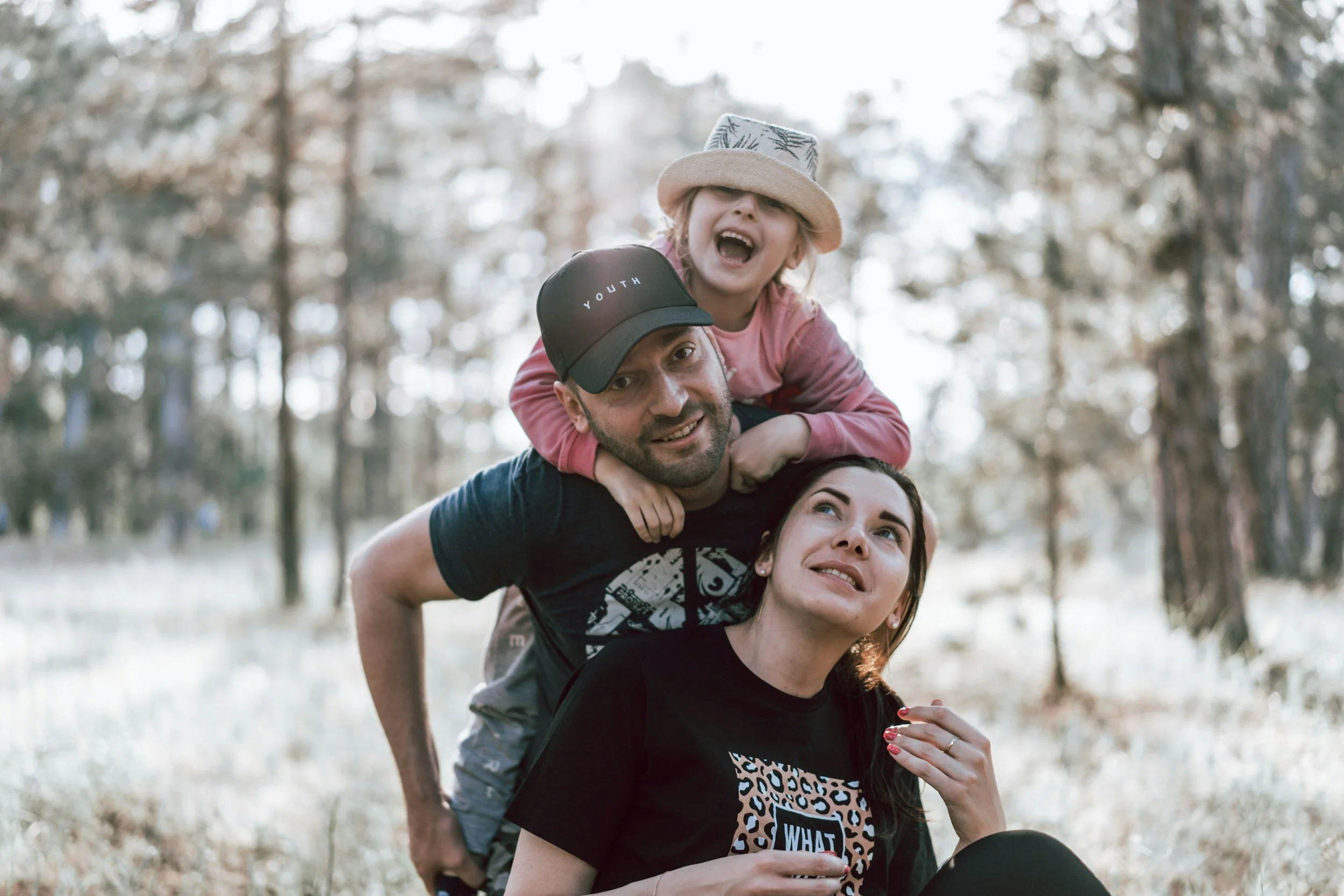 A family enjoying outdoor time in a park, with a man carrying a young girl on his shoulders and a woman sitting on the ground, all smiling and looking happy.