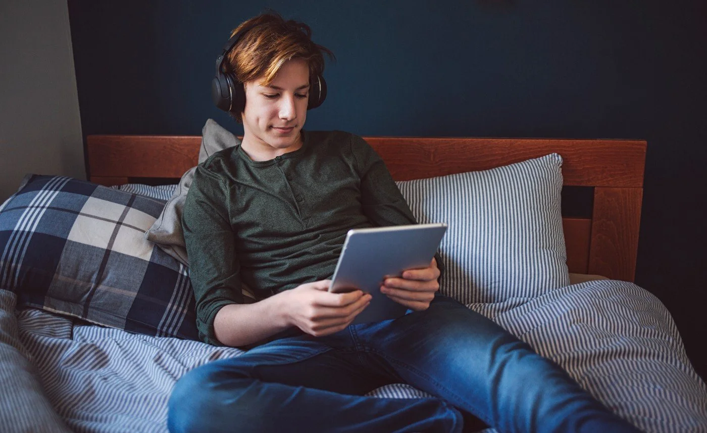 Young person with headphones sitting on bed, looking at a tablet.