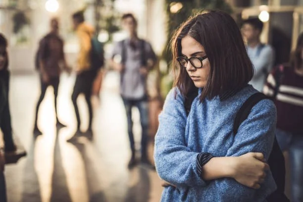 A young woman with dark hair and glasses standing with arms crossed in a public indoor space, appearing upset or contemplative, with blurred people in the background.