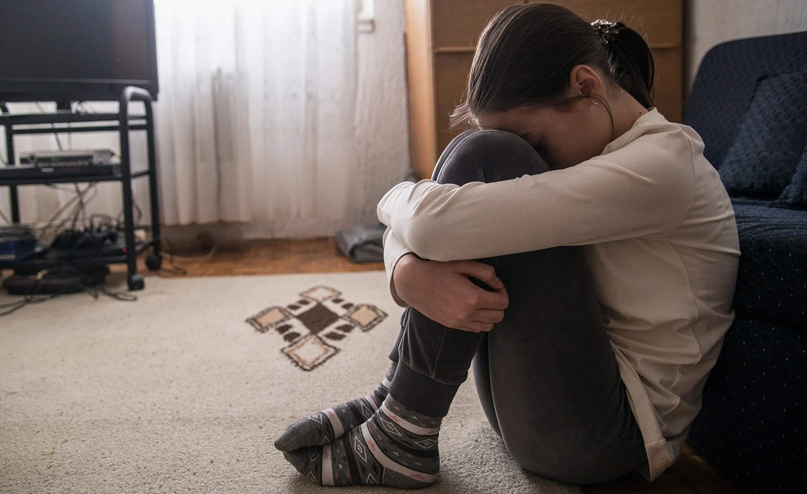 A woman sitting on the floor with her knees drawn up, hugging her knees and resting her forehead on her knees. She appears to be in a distressed or contemplative state, in a room with wooden floors and a beige rug, surrounded by furniture and electronic equipment.