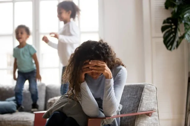 An overwhelmed woman sits on a sofa with her head in her hands while two children play in the background.