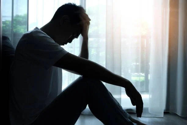 Silhouette of a person sitting on the floor with head in hand near a window with curtains, appearing distressed or sad.