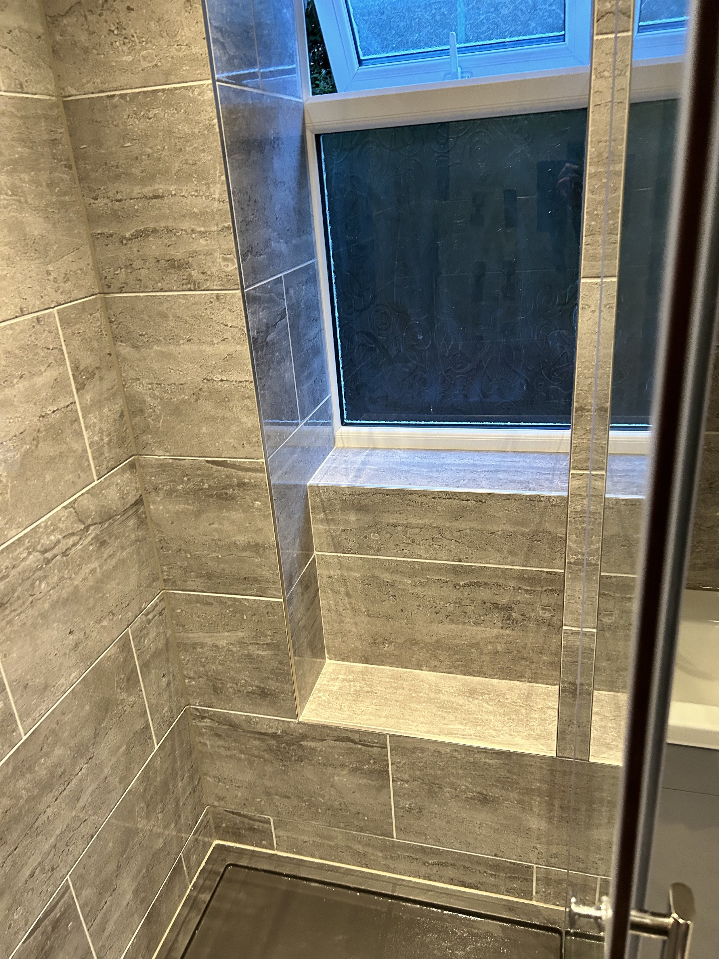 A tiled bathroom shower with beige and gray tiles, a window with frosted glass, and a metal shower drain on the floor.
