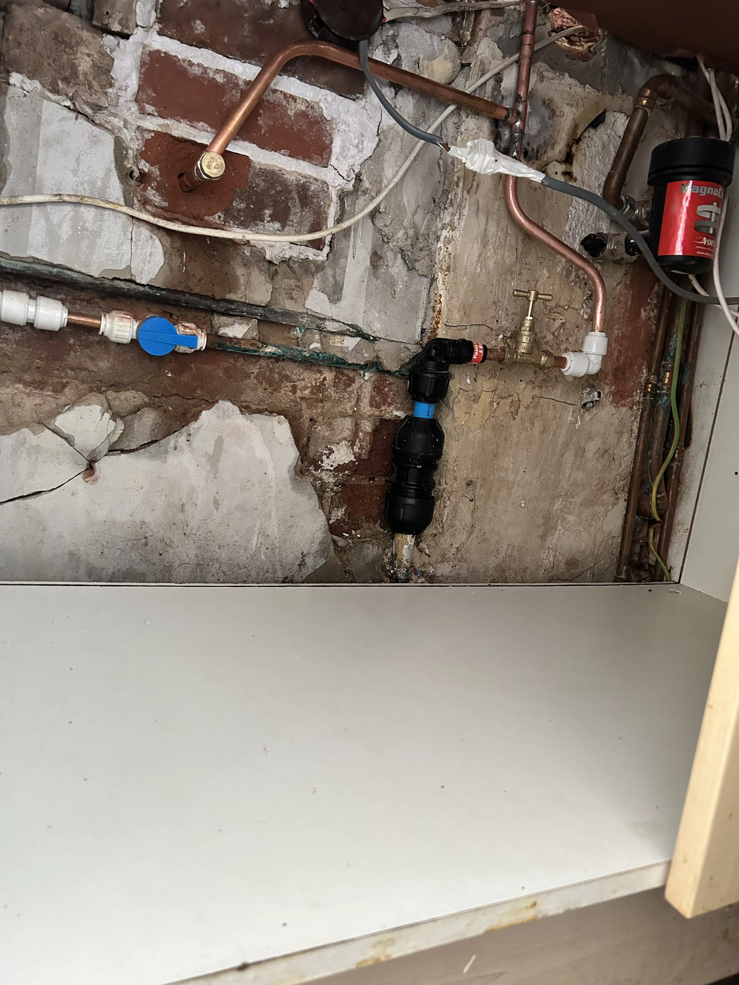 Plumbing pipes and fixtures behind a partially removed wall in a kitchen or utility room, including copper, plastic, and black PEX fittings with a blue valve handle.
