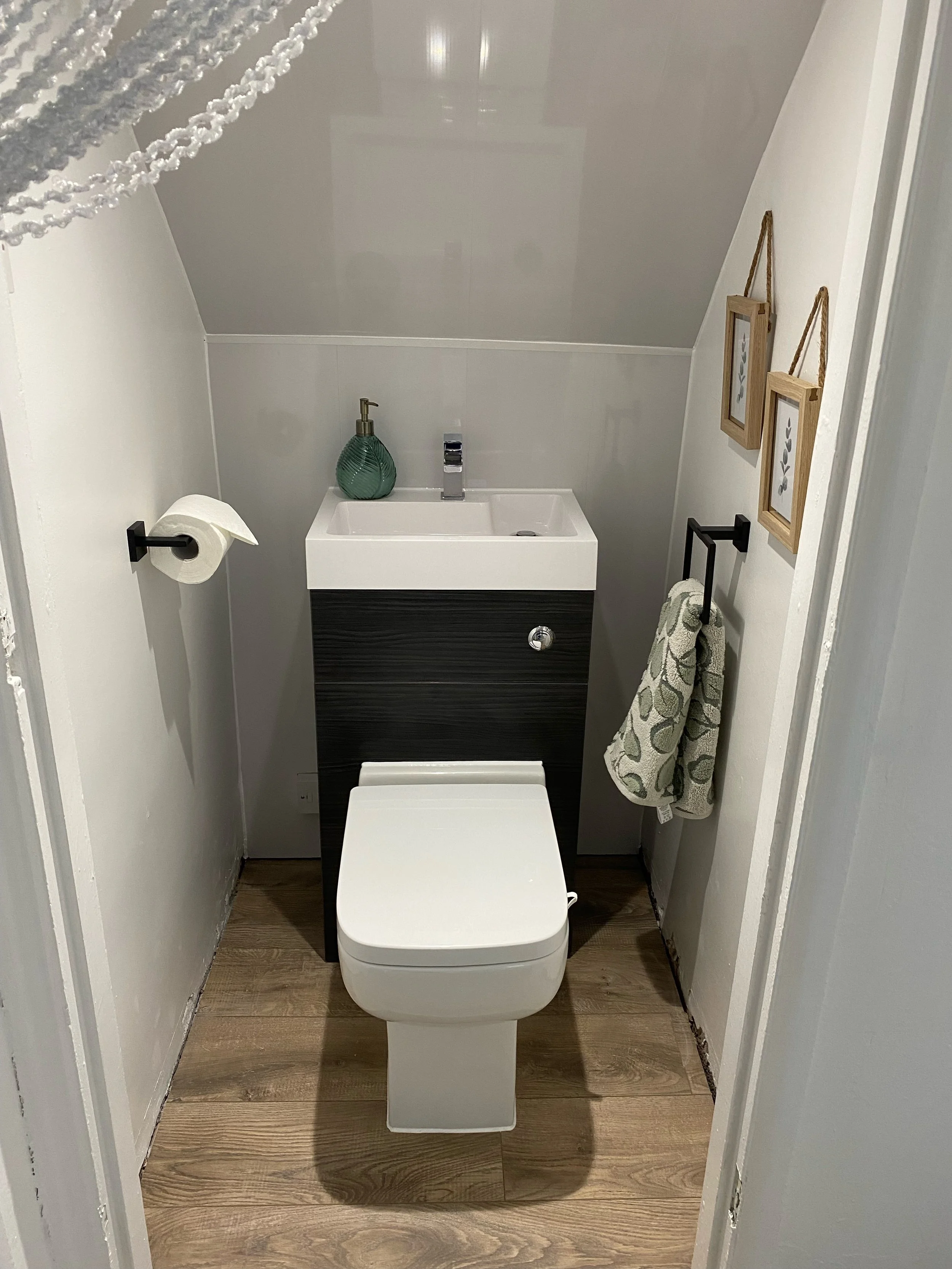 Small modern bathroom with a white toilet, a tiny white sink with a green soap dispenser, wooden floor, and two framed pictures on the wall. A decorative string of beads hangs from the ceiling, and a towel is on a wall-mounted towel rack.