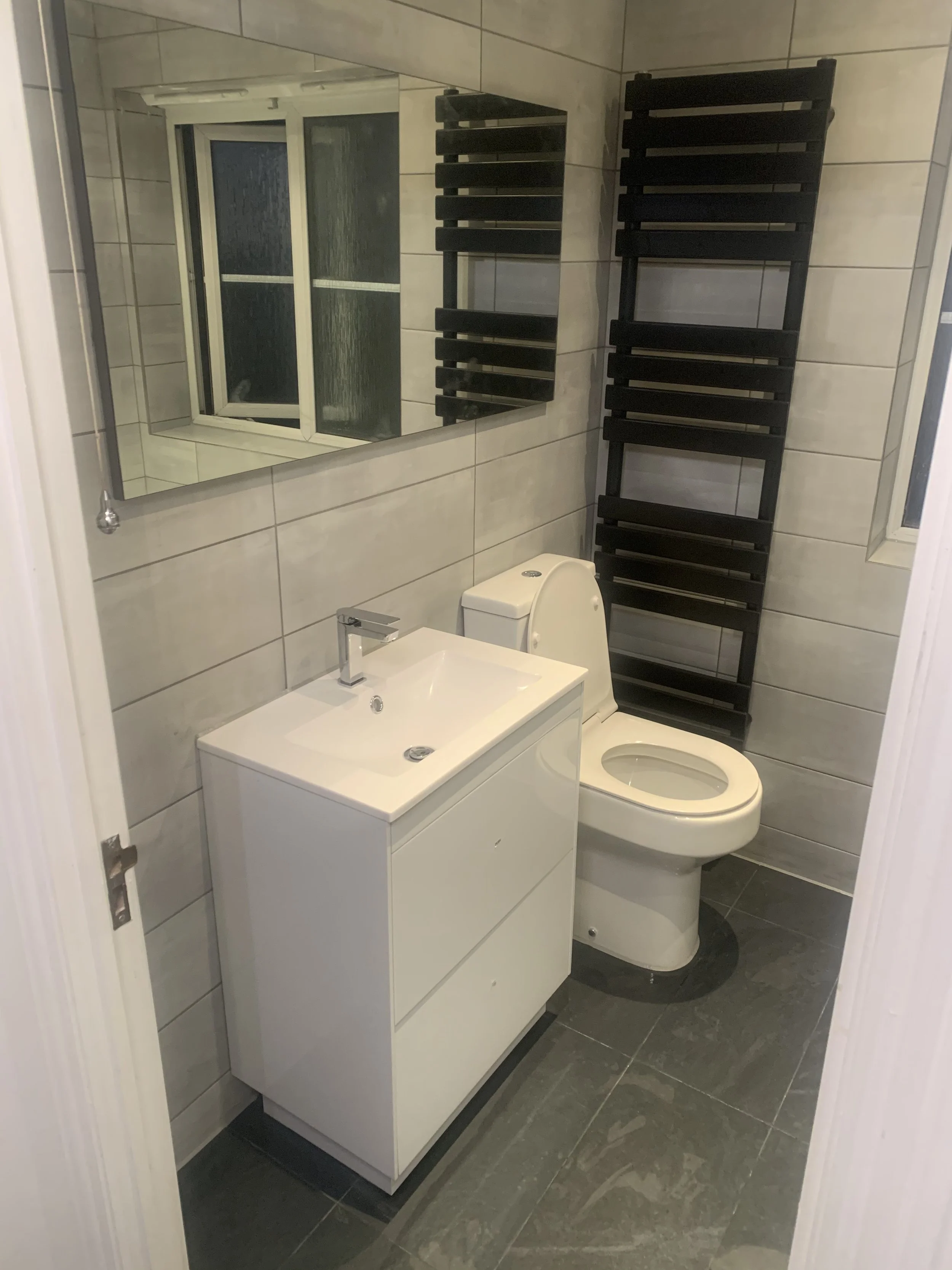 Small bathroom with a white sink vanity, a toilet, and a black heated towel rack. A large mirror hangs above the sink, and there is a window with frosted glass and sliding panels.