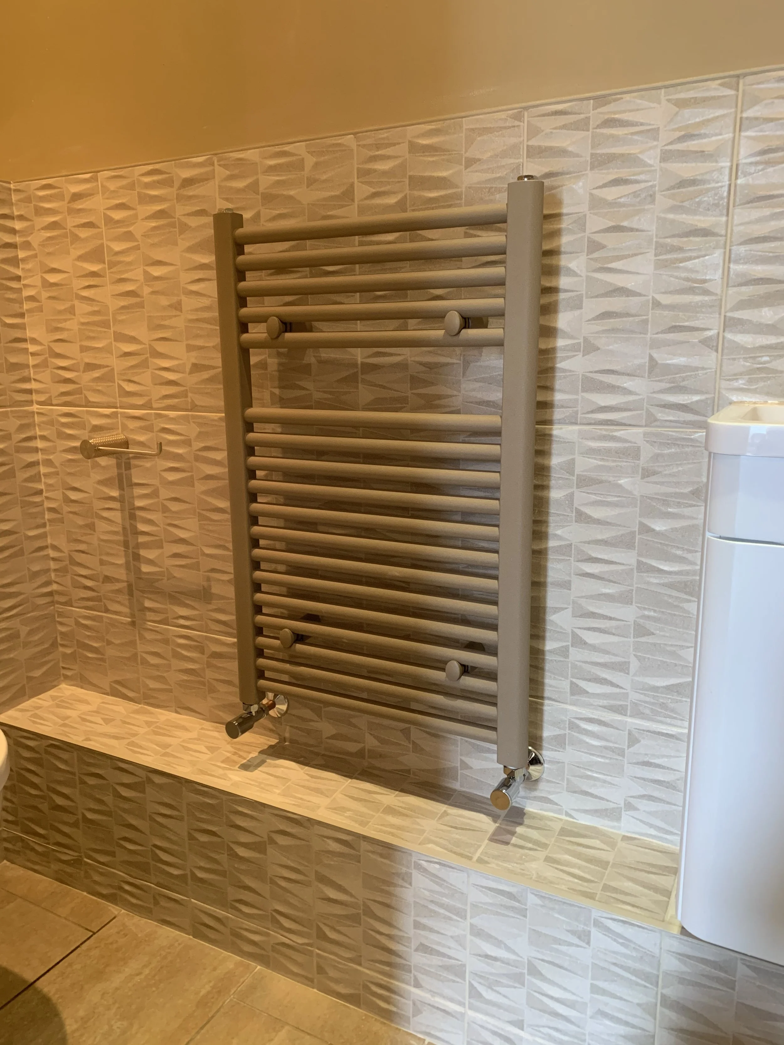 A wall-mounted heated towel rack in a bathroom with beige tiled walls.