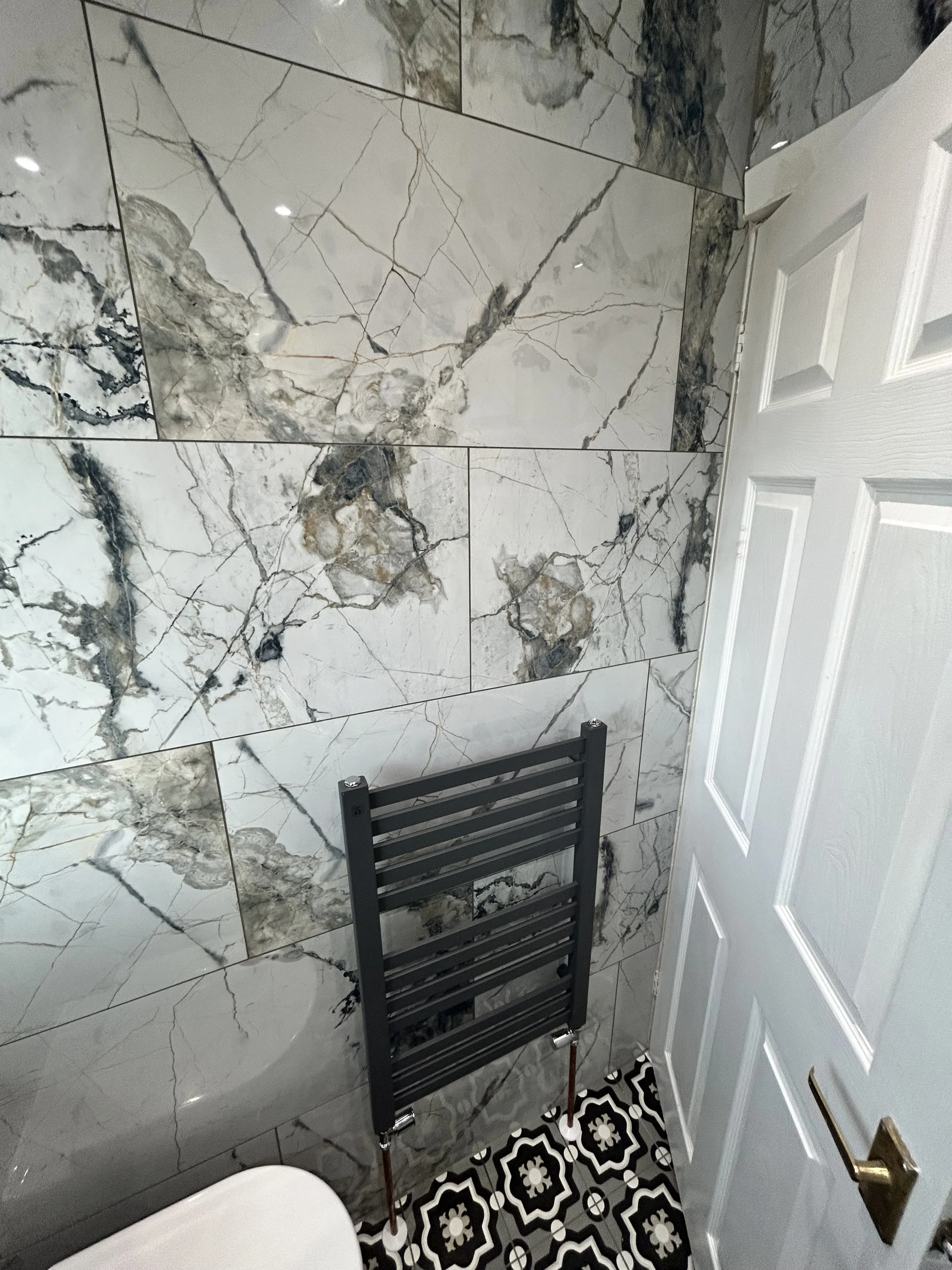 A small bathroom featuring large marble tiles with gray and black veining on the wall, a patterned black-and-white floor, a white door, and a black heated towel rack.