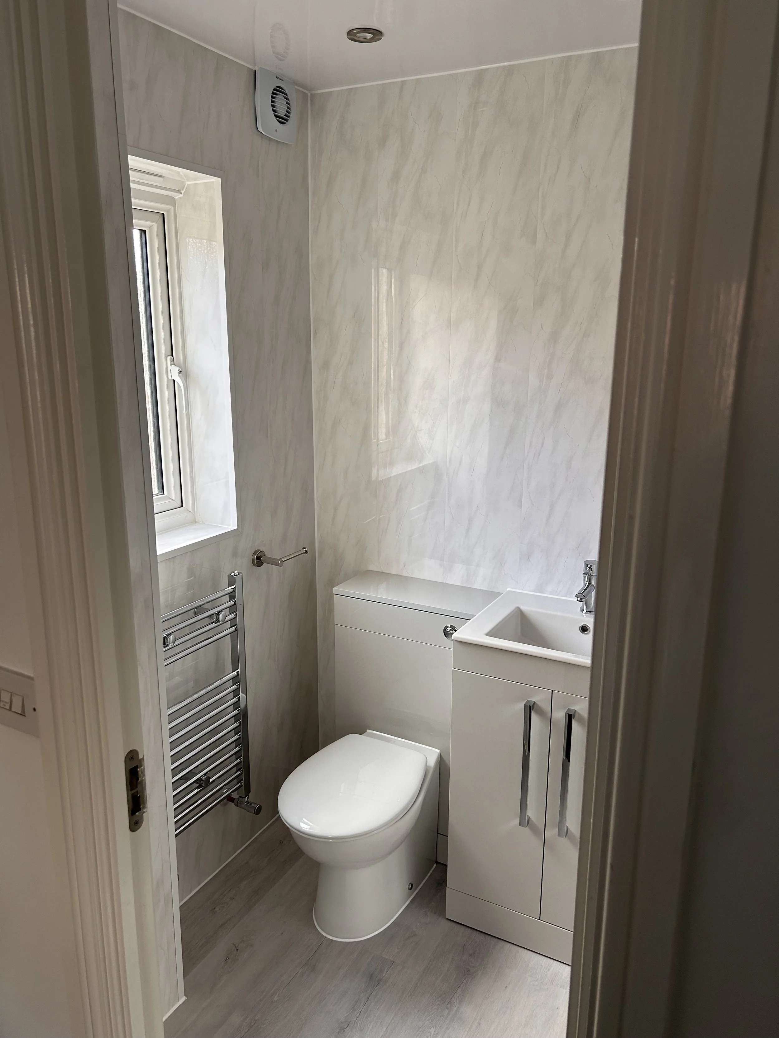 Small bathroom with white toilet, white sink with cabinet, small window, beige walls, wooden flooring, and a towel rack.