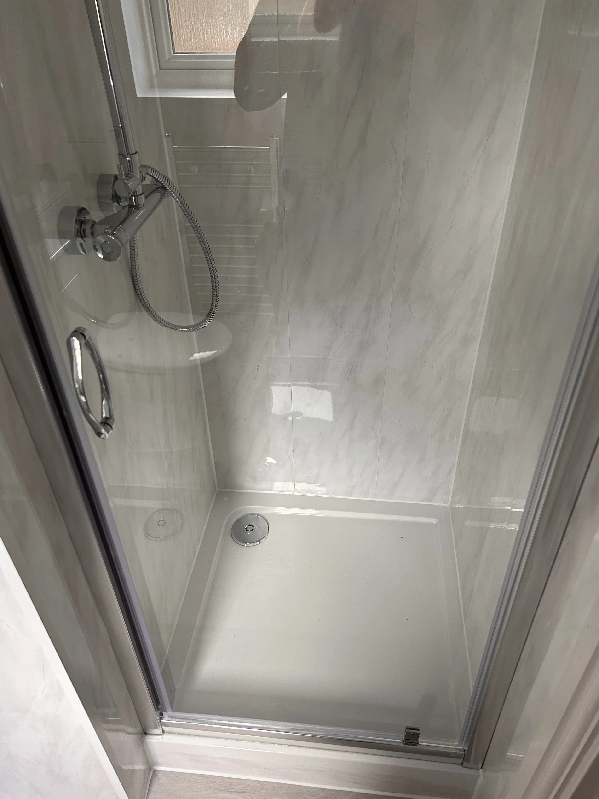 Empty glass shower stall with a chrome handheld showerhead and sliding door, with a tiled marble wall and a drain on the shower floor.