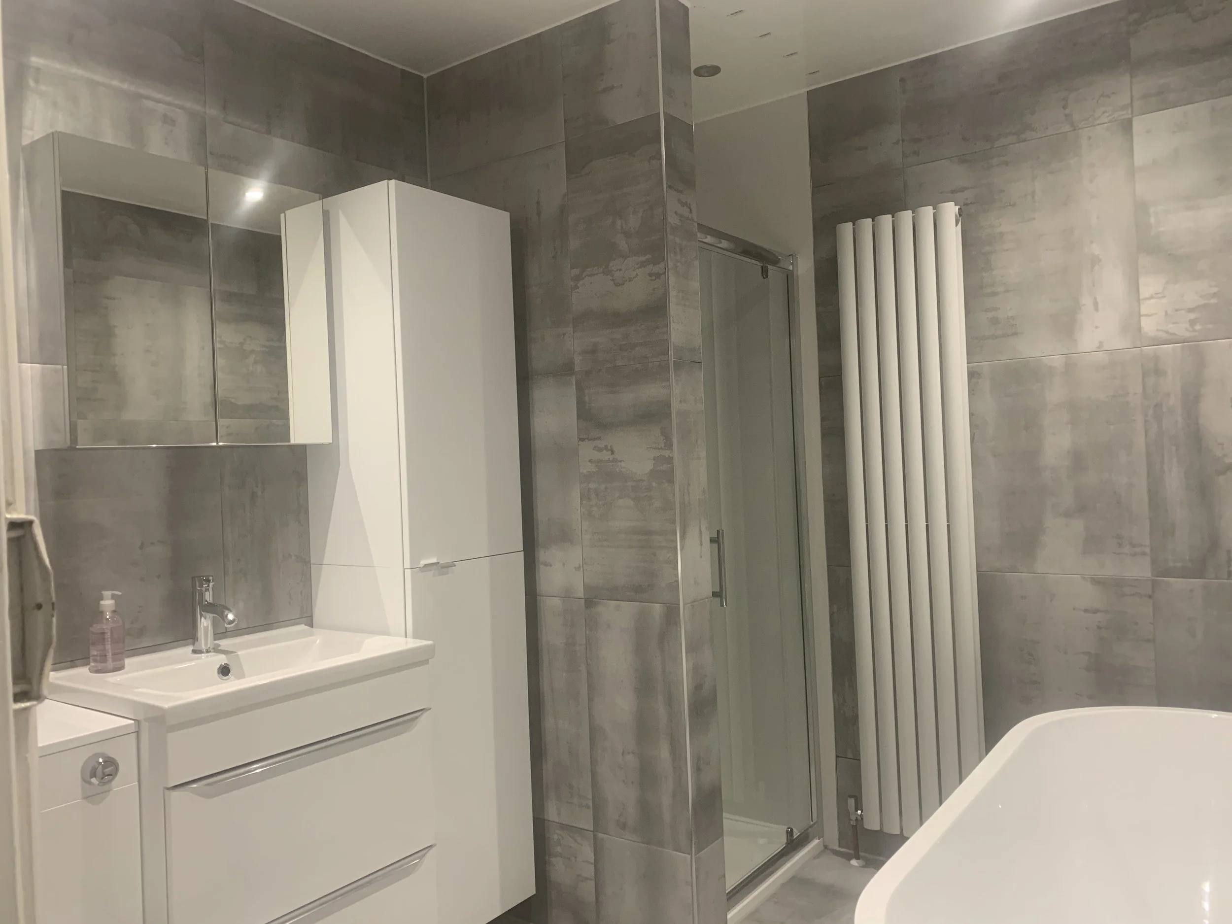 A modern bathroom with gray tiled walls, a white vanity with a sink and a mirror cabinet, a tall white storage cabinet, a glass shower enclosure, a white vertical towel warmer, and a bathtub.