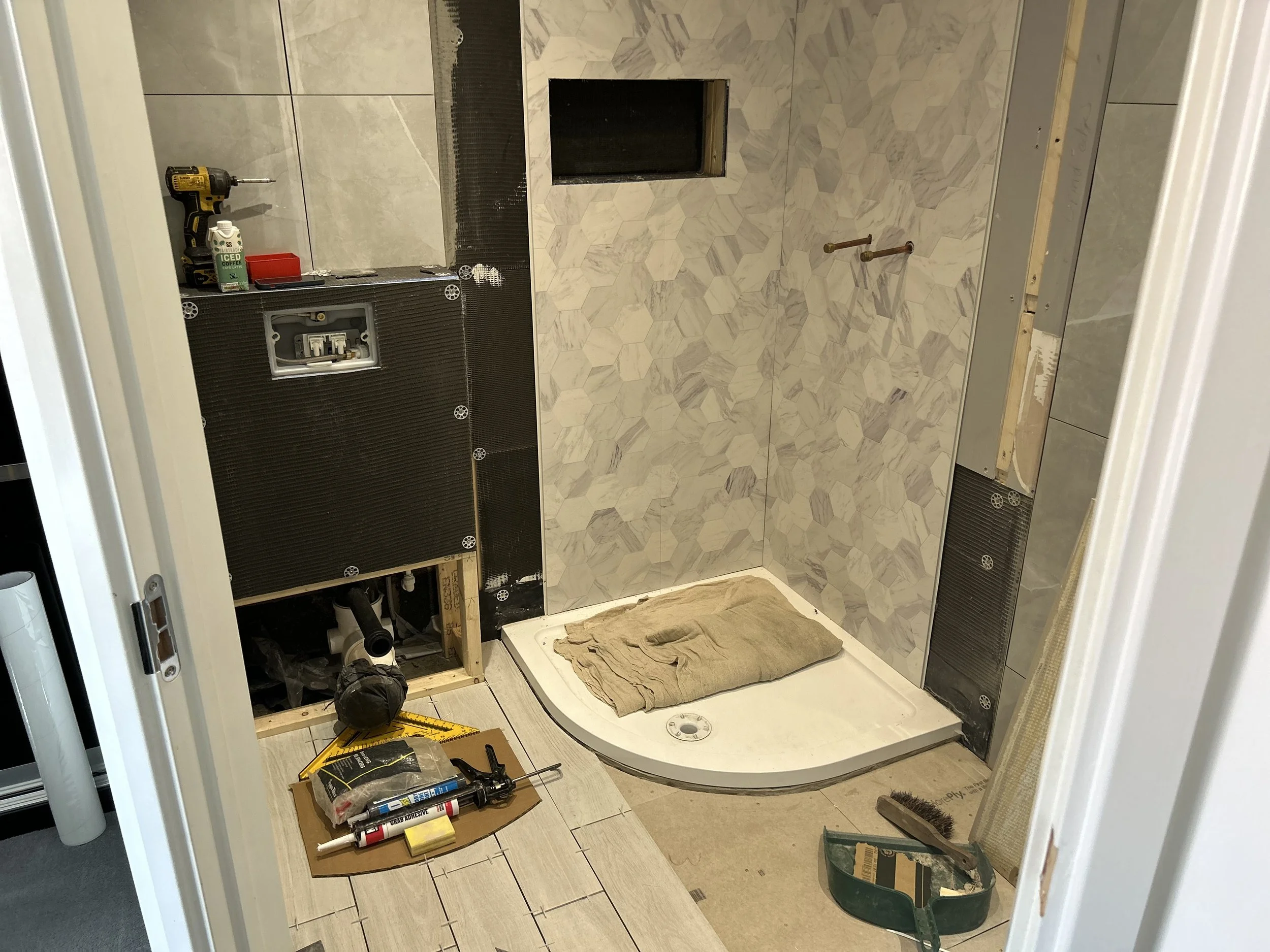 Bathroom under renovation showing a shower base, partial wall tiling, construction tools, and materials.