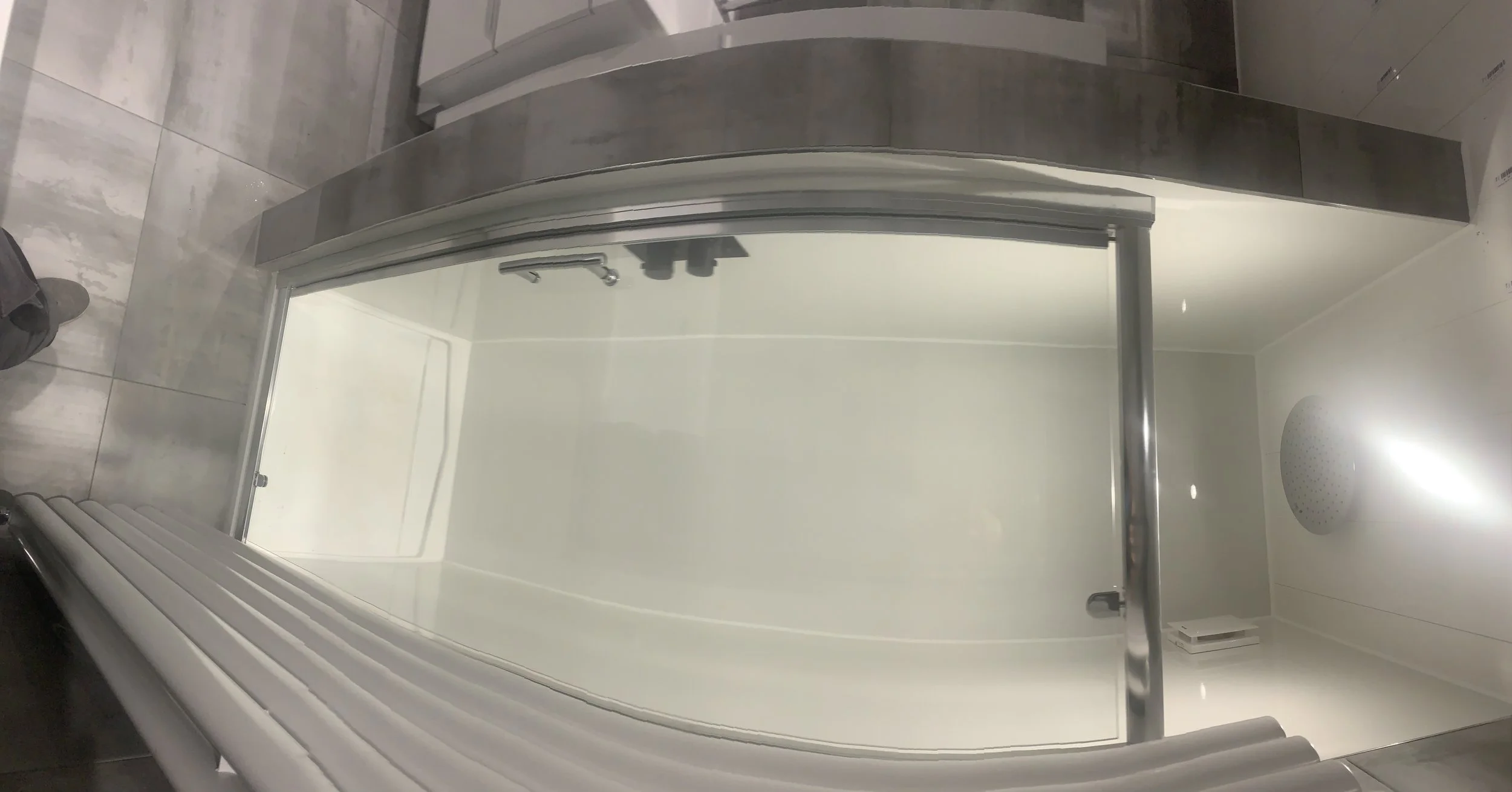 Empty shower stall with sliding glass door, silver frame, white walls, and overhead showerhead, located in a modern bathroom with gray tiled floor.