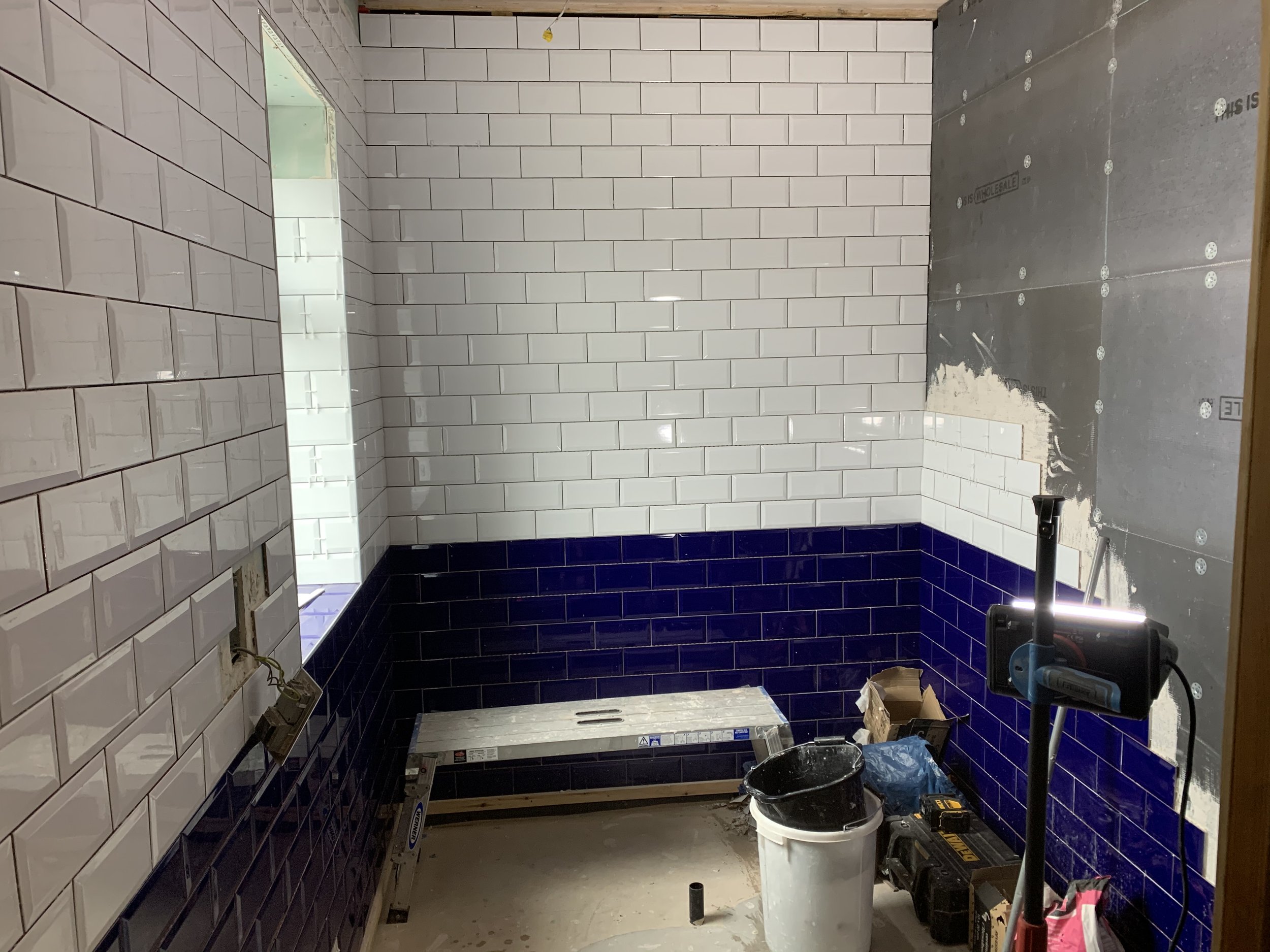 A room under renovation with white subway tiles partially installed on walls, a ladder on the floor, construction tools, a bucket, and paint supplies.