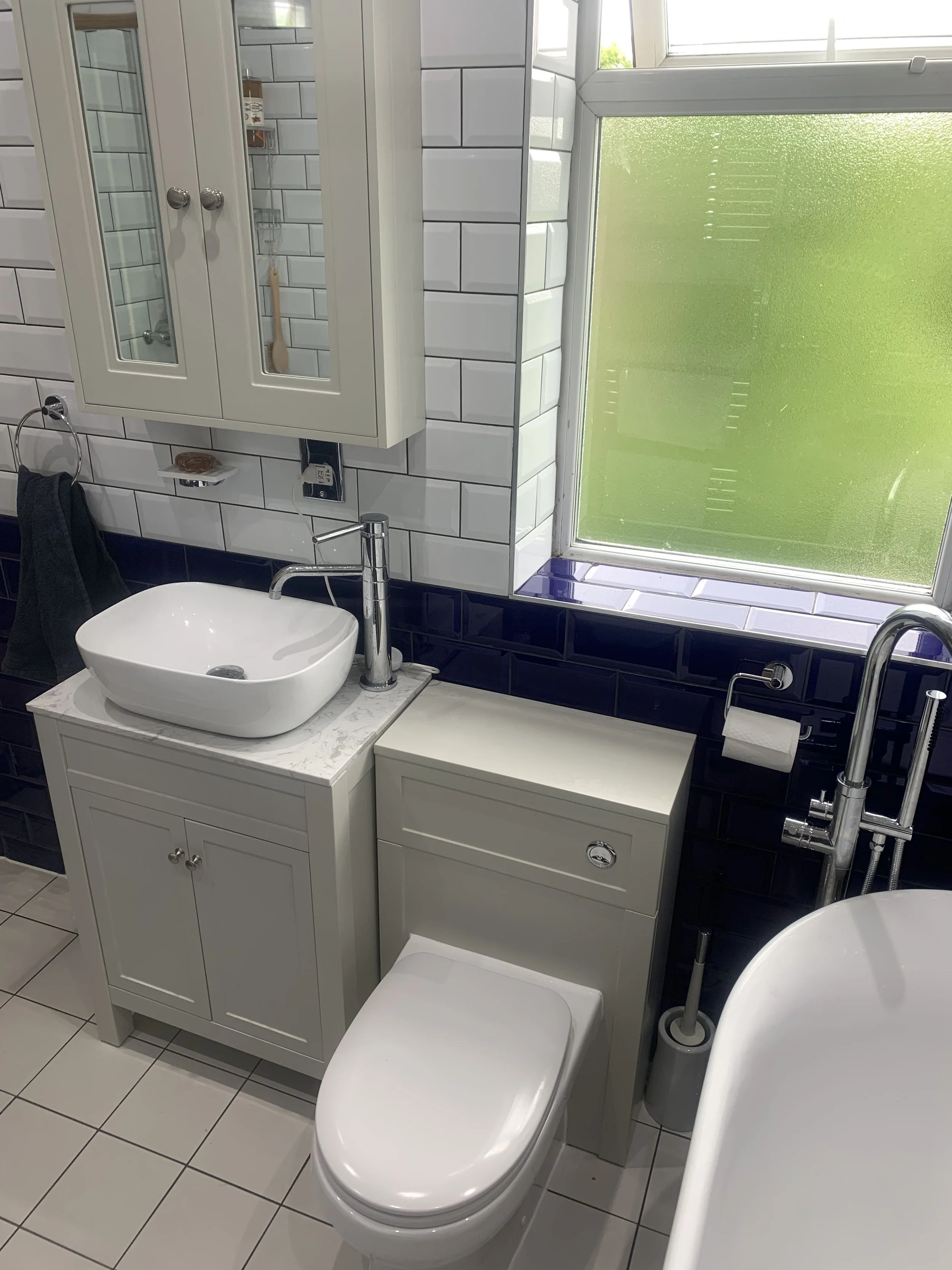 A small bathroom with white and dark blue tile walls, a white vanity with a vessel sink and chrome faucet, a mirrored cabinet, a frosted window, and a white toilet.