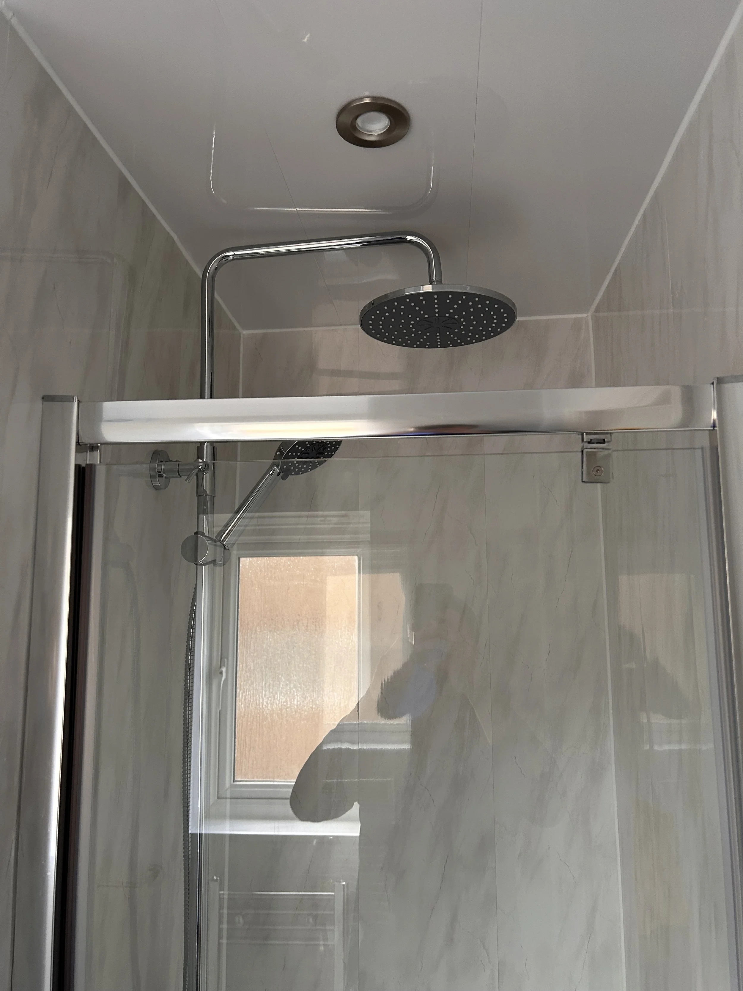 A bathroom shower with a rain shower head and a handheld shower head, enclosed by a glass door, with a small window and a mirror reflecting a person taking the photo.