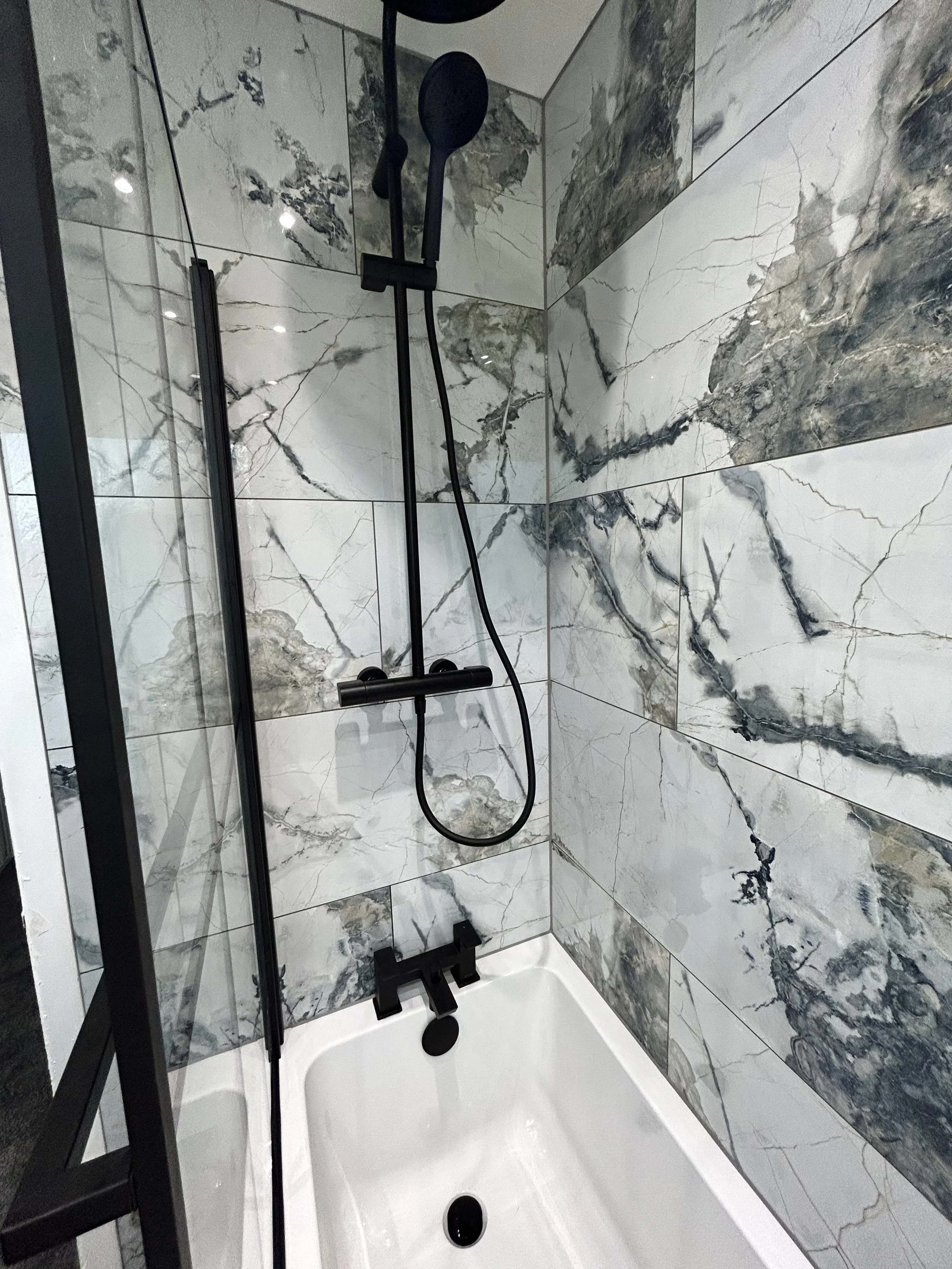 A modern shower with marble-patterned tiles and black fixtures, including a handheld showerhead and a glass enclosure.