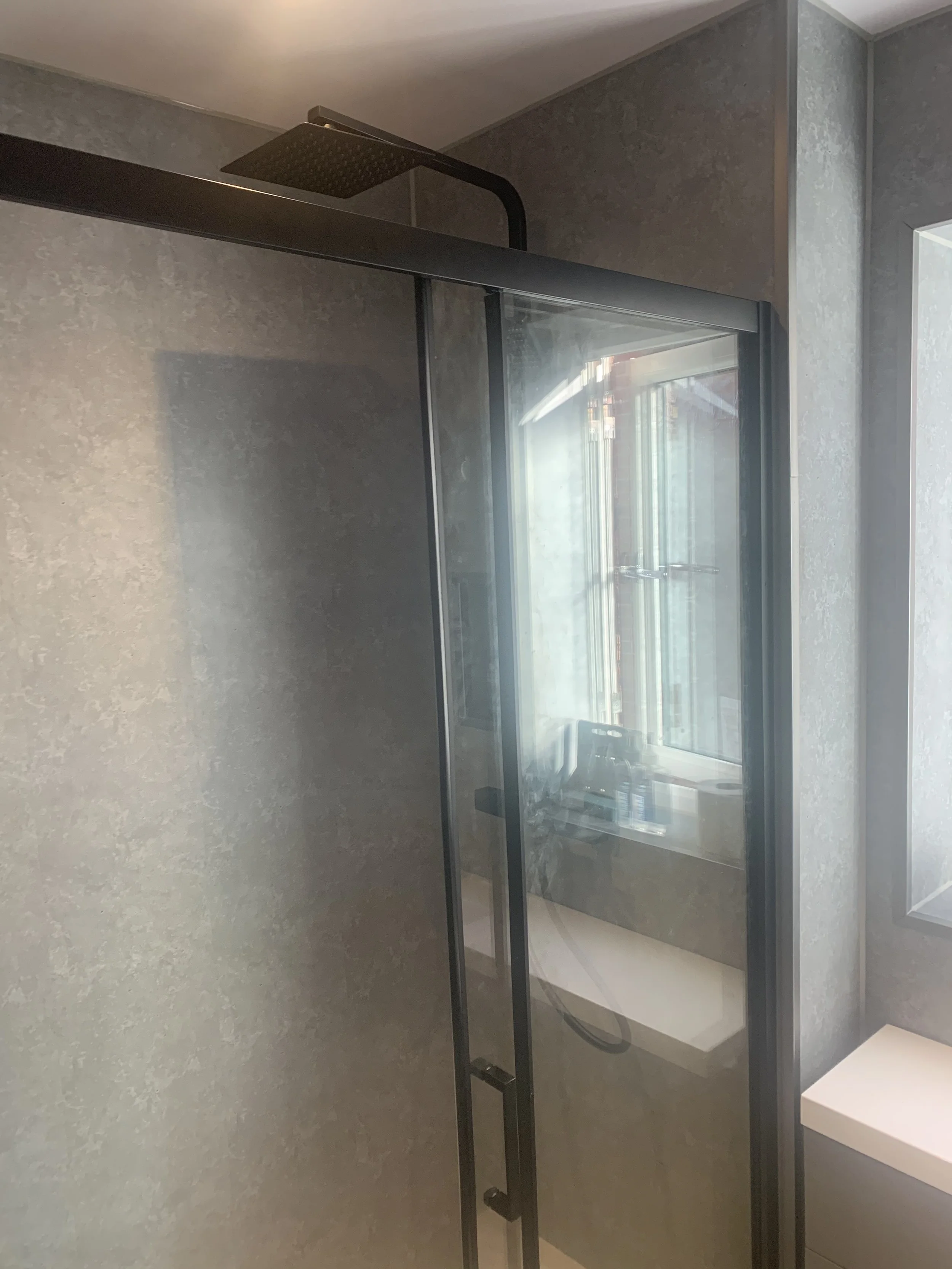 Modern shower with a black frame, glass door, black showerhead, and a window in a bathroom with gray textured walls.