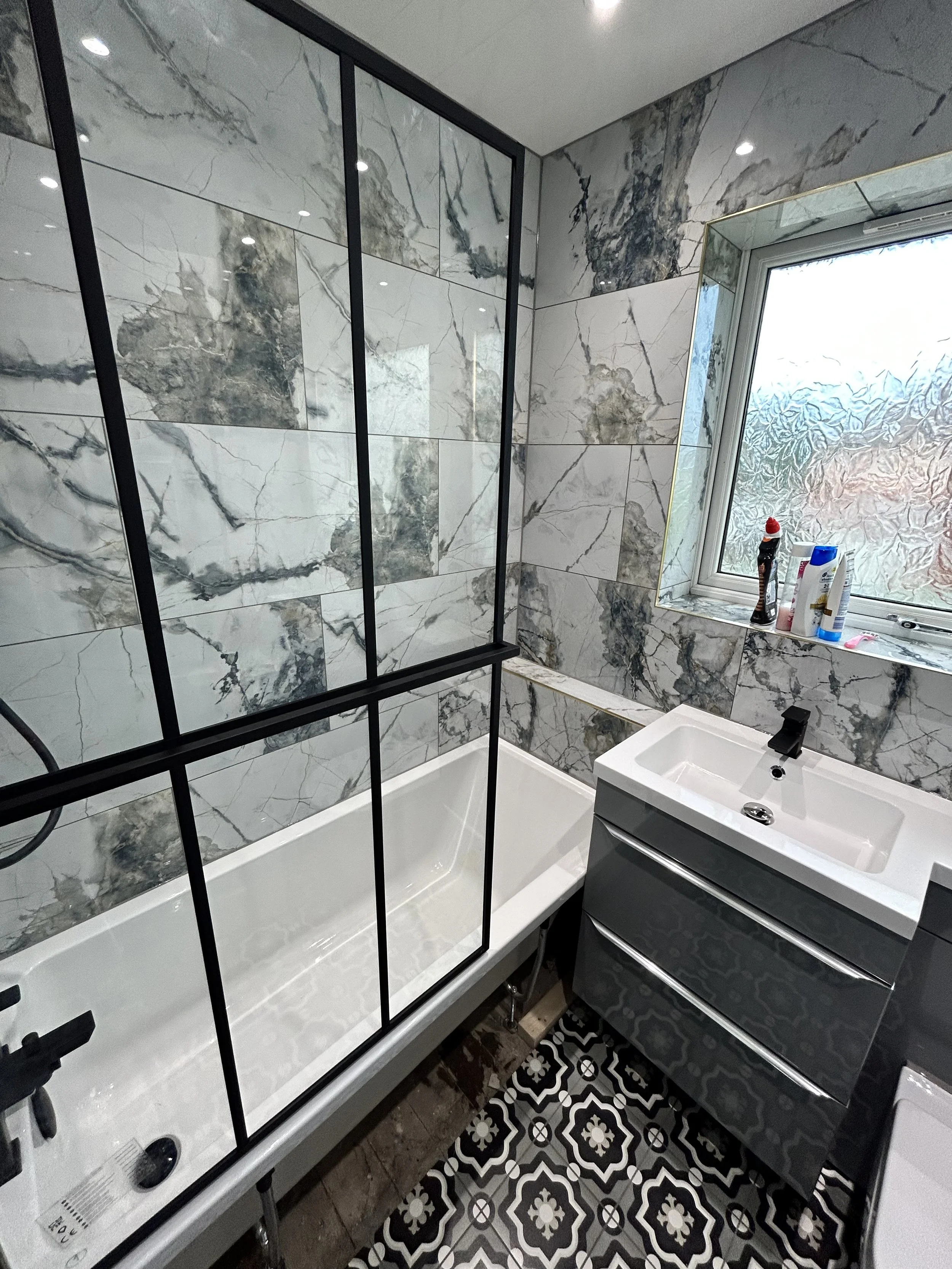 Bathroom with marble tiled walls, a bathtub with a black metal framed glass shower screen, a small gray vanity with a white sink, and a window on the right with cleaning supplies on the sill. The floor has patterned black and white tiles.