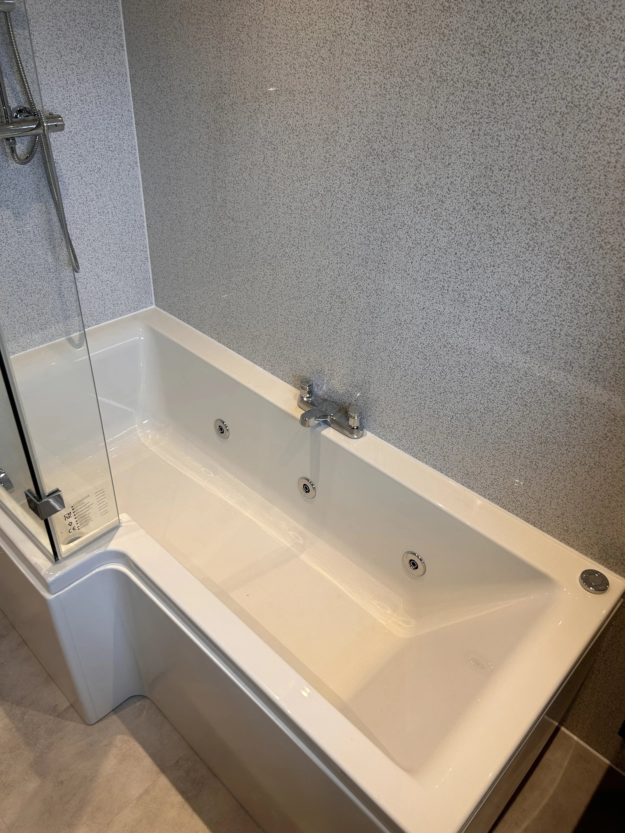 A white rectangular bathtub with four jets is situated against a textured gray wall, with a faucet mounted on the wall above the tub. There's a glass shower partition on the left side of the bathtub.