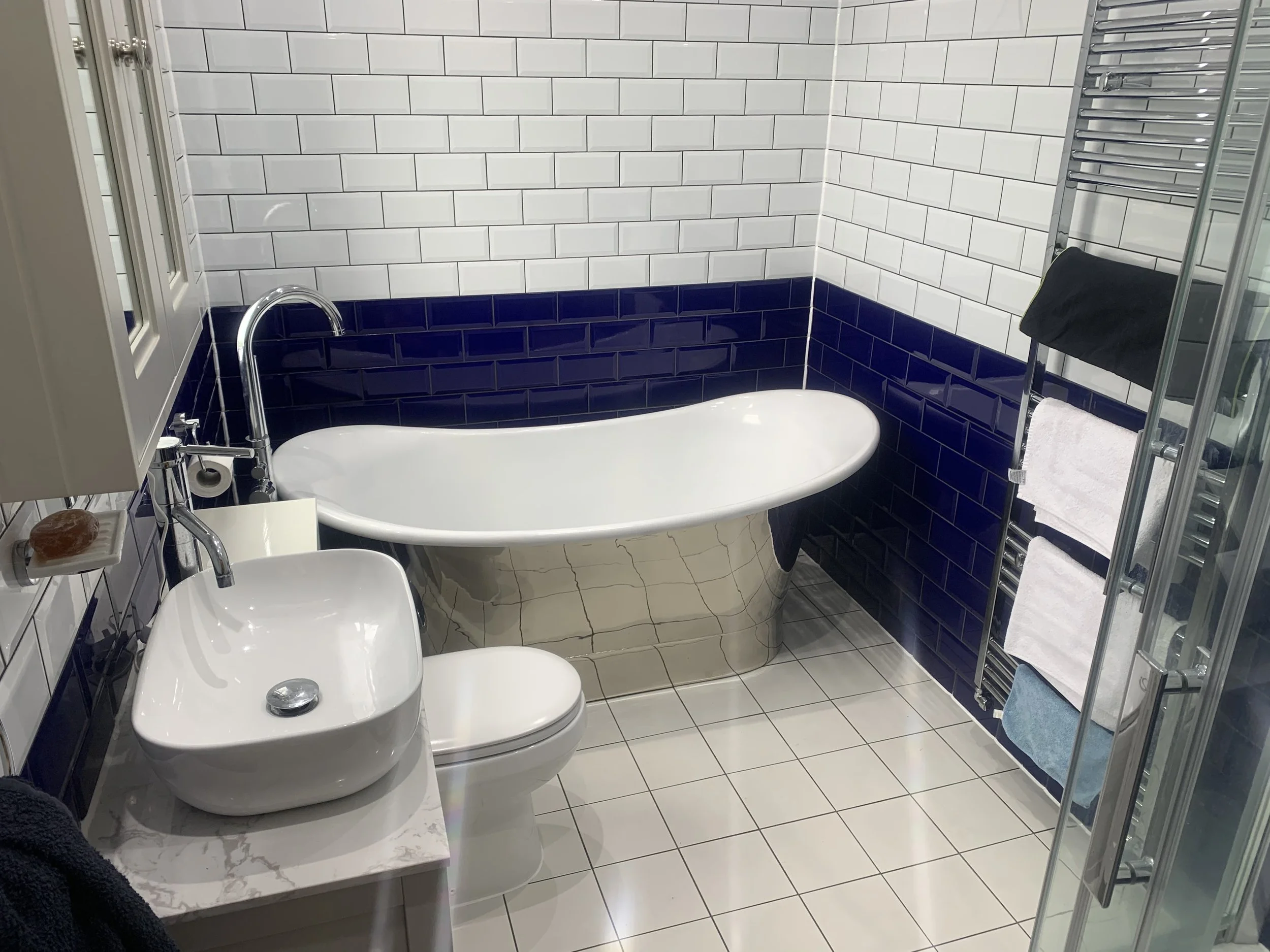 Bathroom with a white freestanding bathtub, a sink with a marble top, a toilet, a towel rack with towels, and tiled walls and floors.