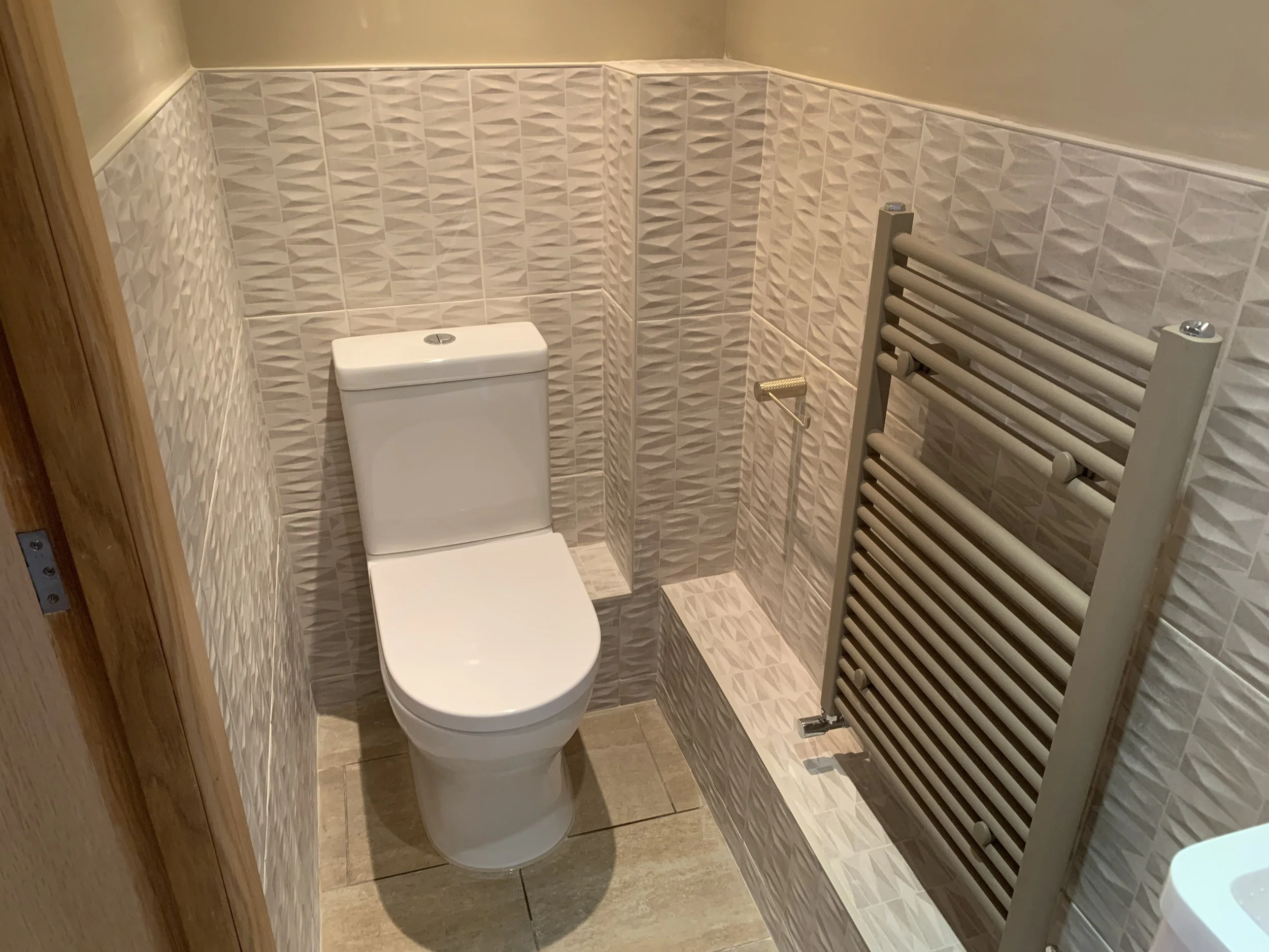 A small bathroom with a white toilet, beige textured wall tiles, and a gray towel warmer radiator.