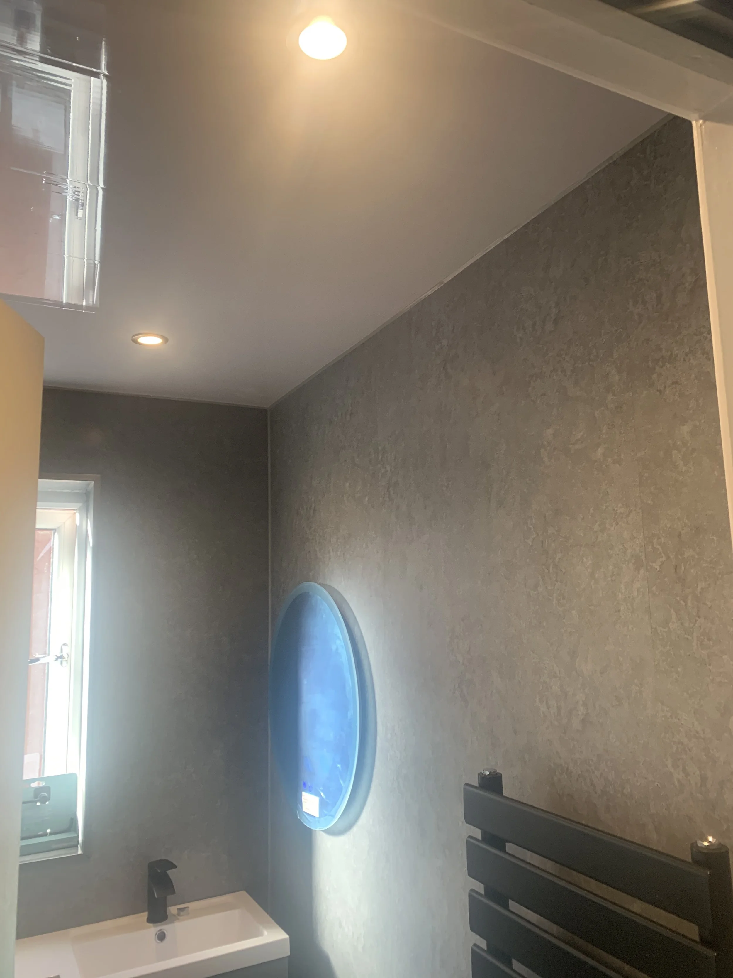 Modern bathroom with gray textured walls, a small sink, a black heated towel rack, a window, and a blue circular mirror.