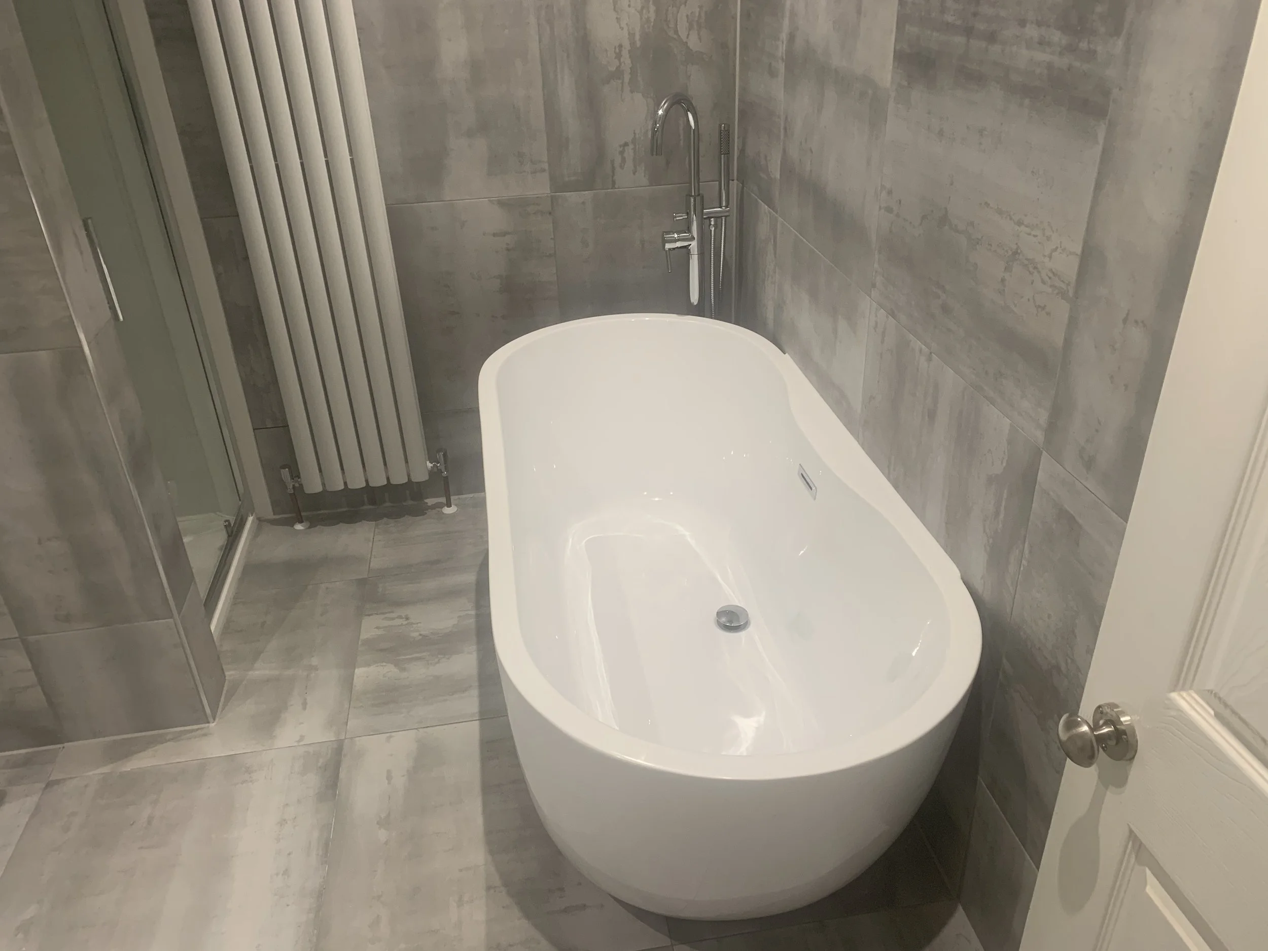 Modern bathroom with freestanding bathtub, tiled walls and floor, a heated towel rail, a shower enclosure, and a white door.