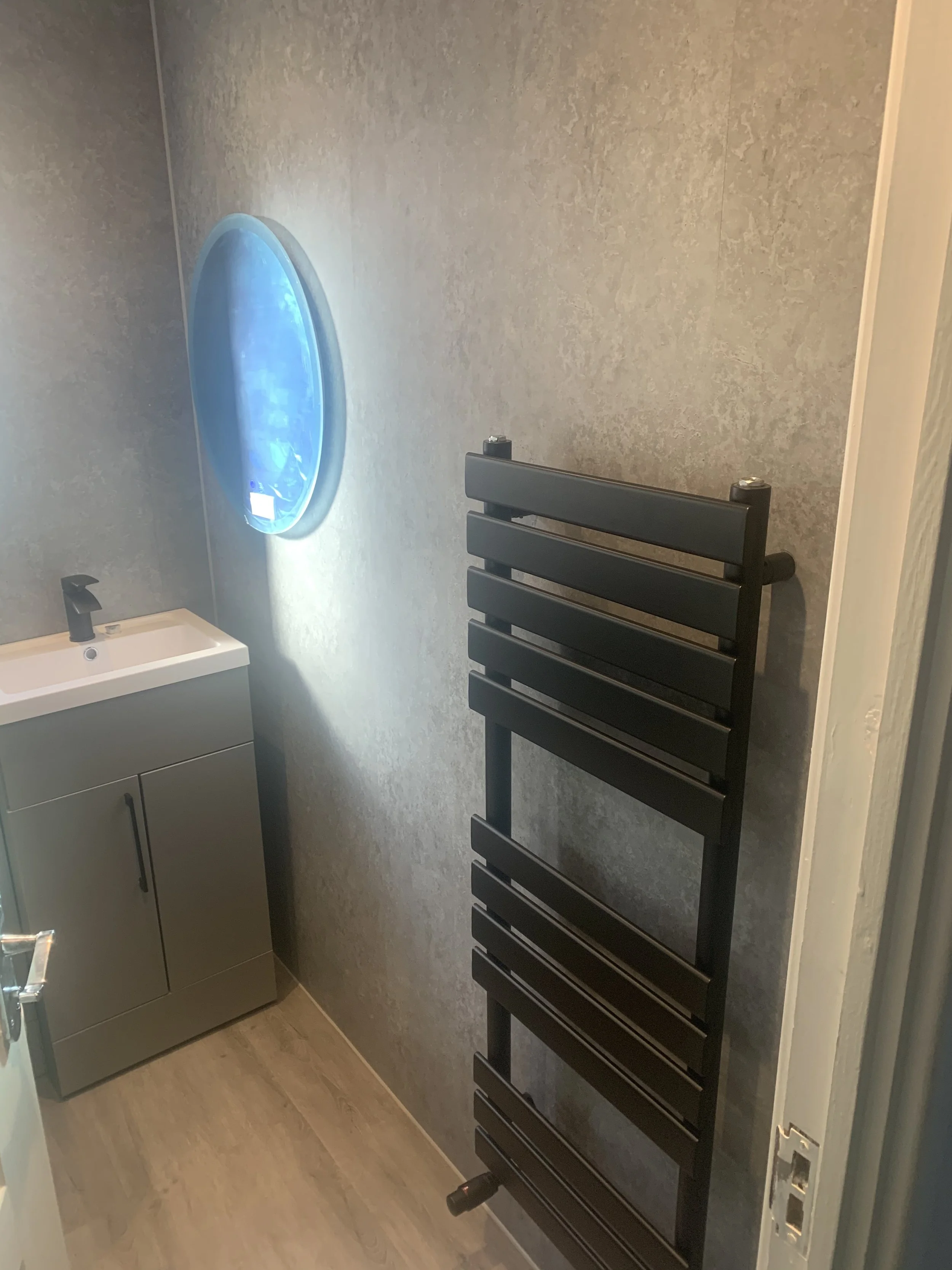 Small bathroom with a modern sink and faucet, a black heated towel rack, and a circular mirror with built-in LED lighting.