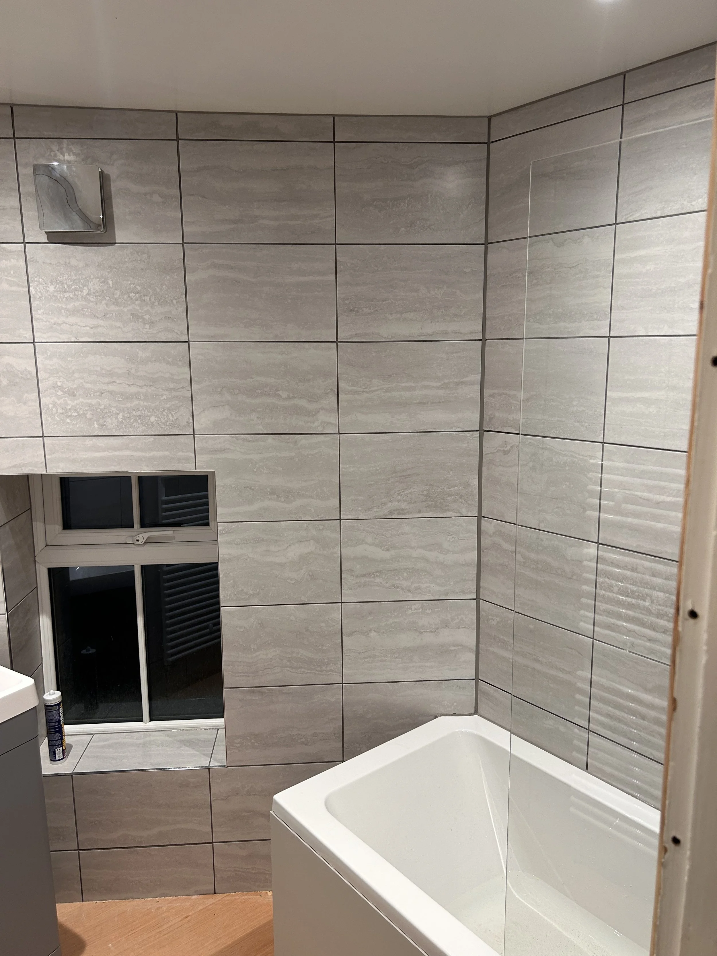 Bathroom with gray tiled walls, a small window, a white bathtub, and a loudspeaker in the upper left corner.