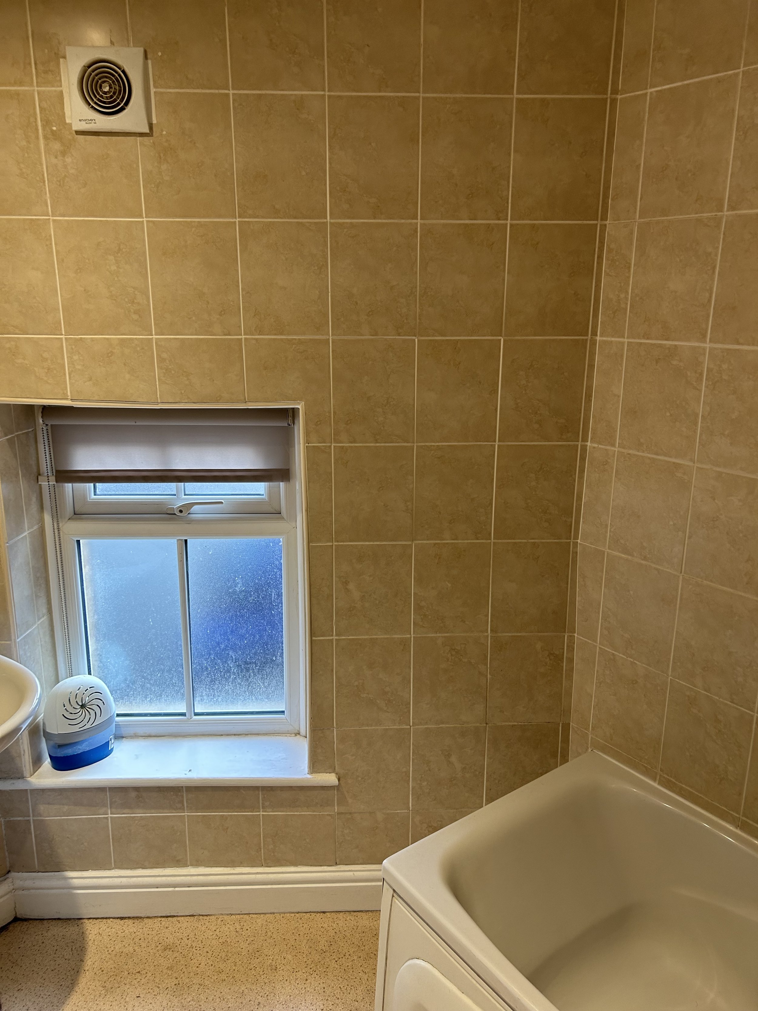 A small bathroom with beige tiled walls, a window with a blind, a white sink, and a partial view of a washing machine.