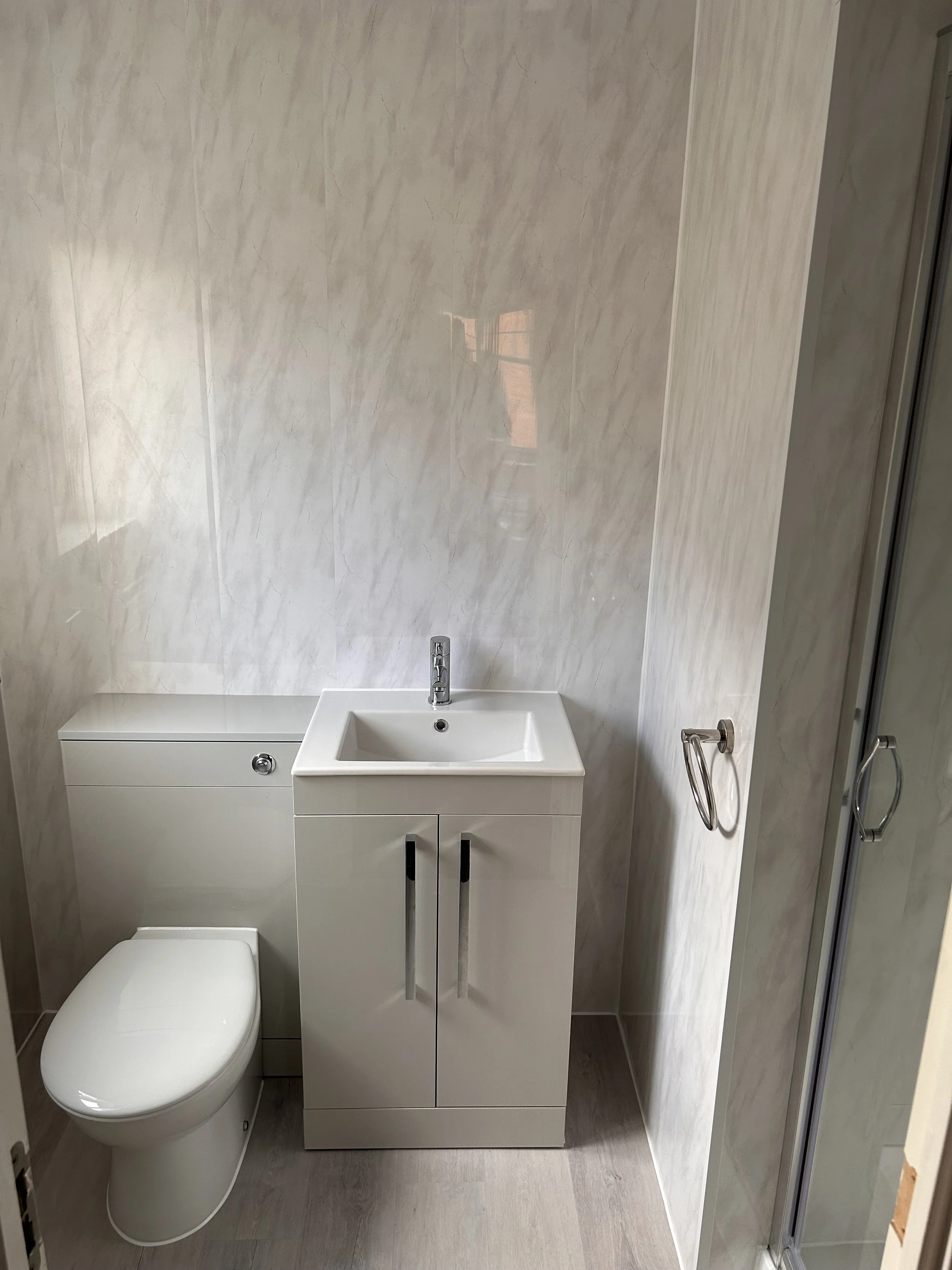 Small modern bathroom with white toilet, white sink in cabinet, and a clear glass shower door on the right; beige marble-like wall and light wood flooring.