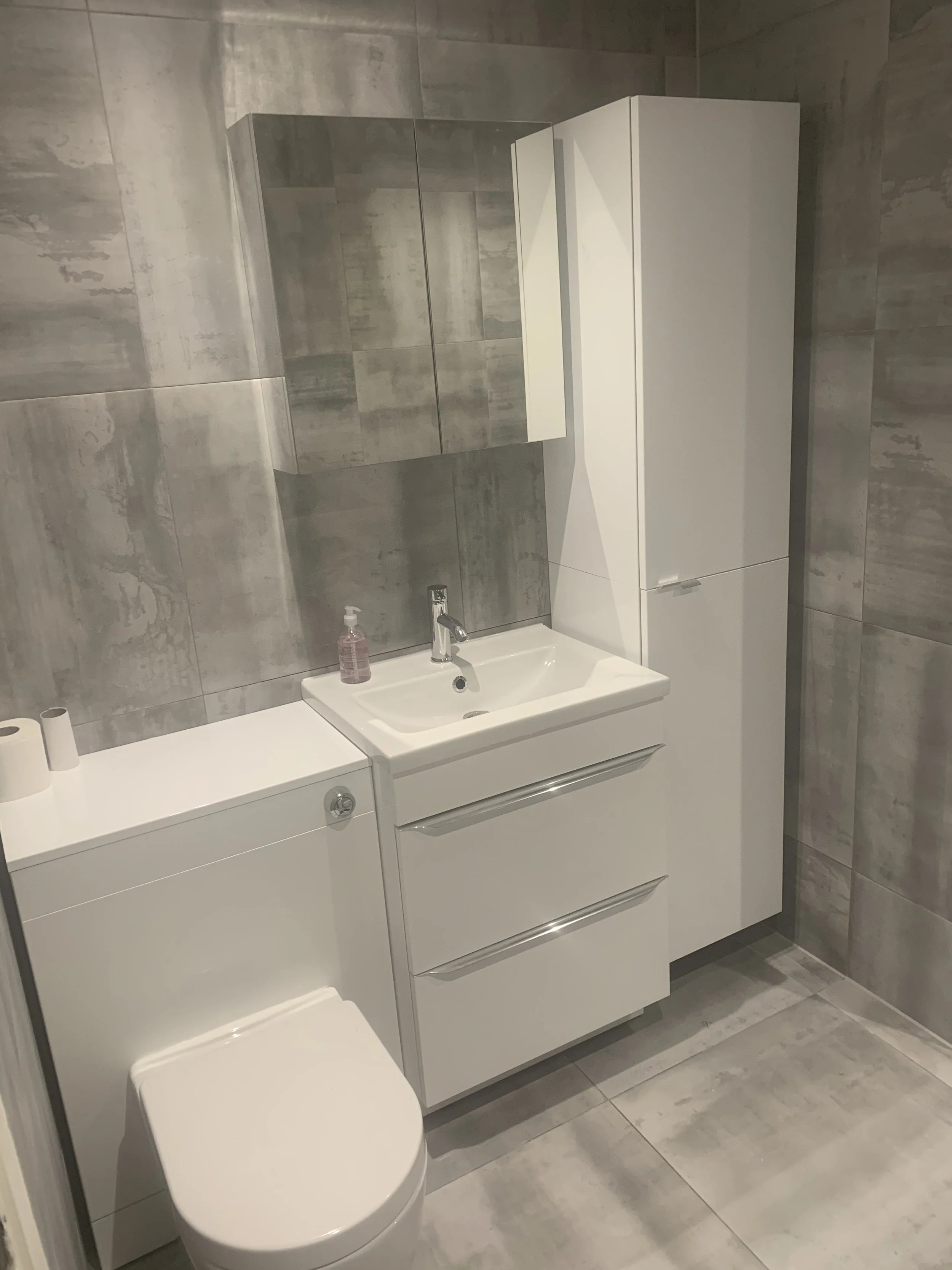 Clean modern bathroom with gray tiled walls, a white toilet, a white vanity with a sink, a mirror cabinet, and a tall white storage cabinet.