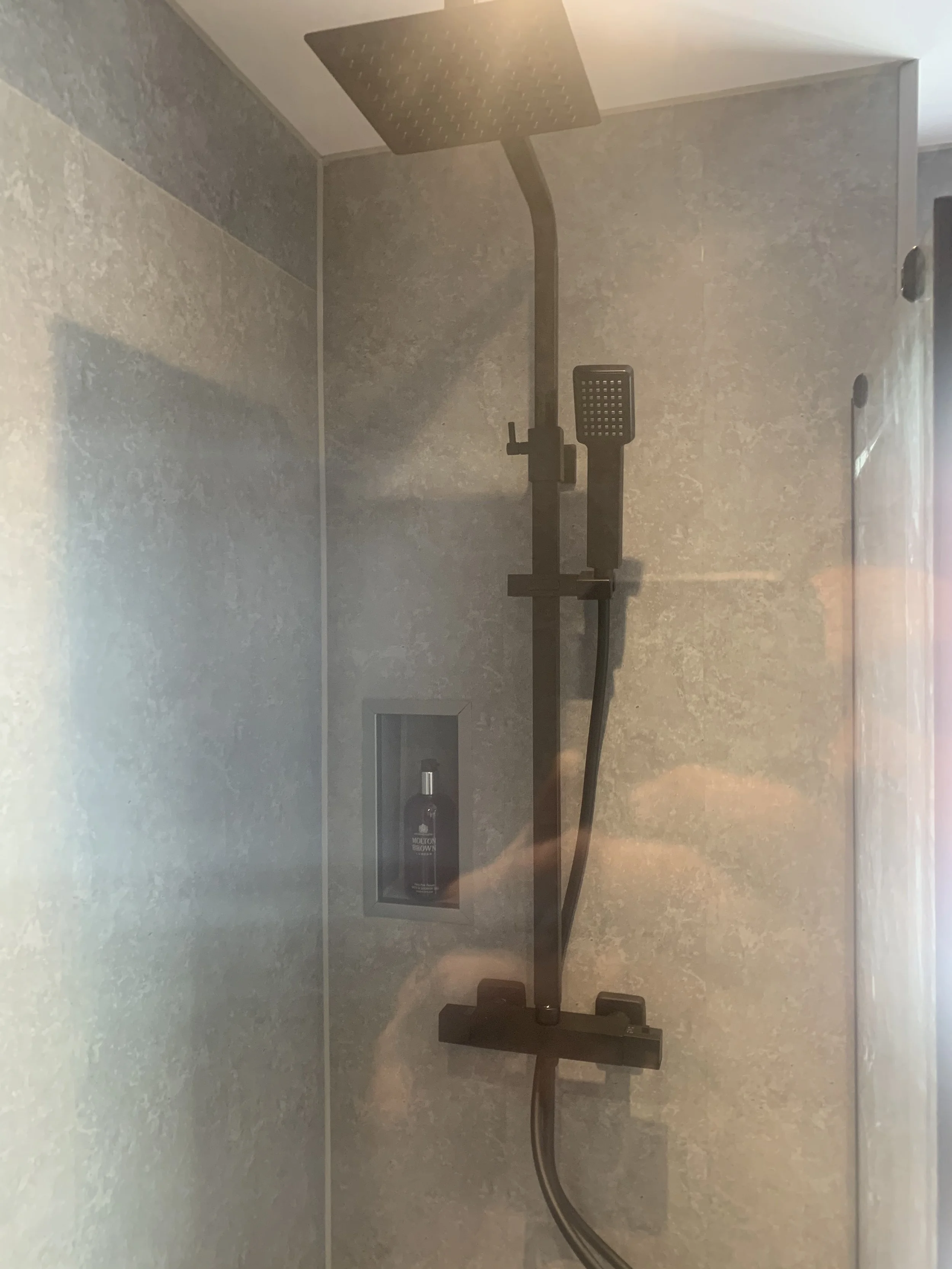Shower with black rainfall shower head, handheld shower, and built-in shelf with a bottle of Molton Brown soap on a gray tiled wall.