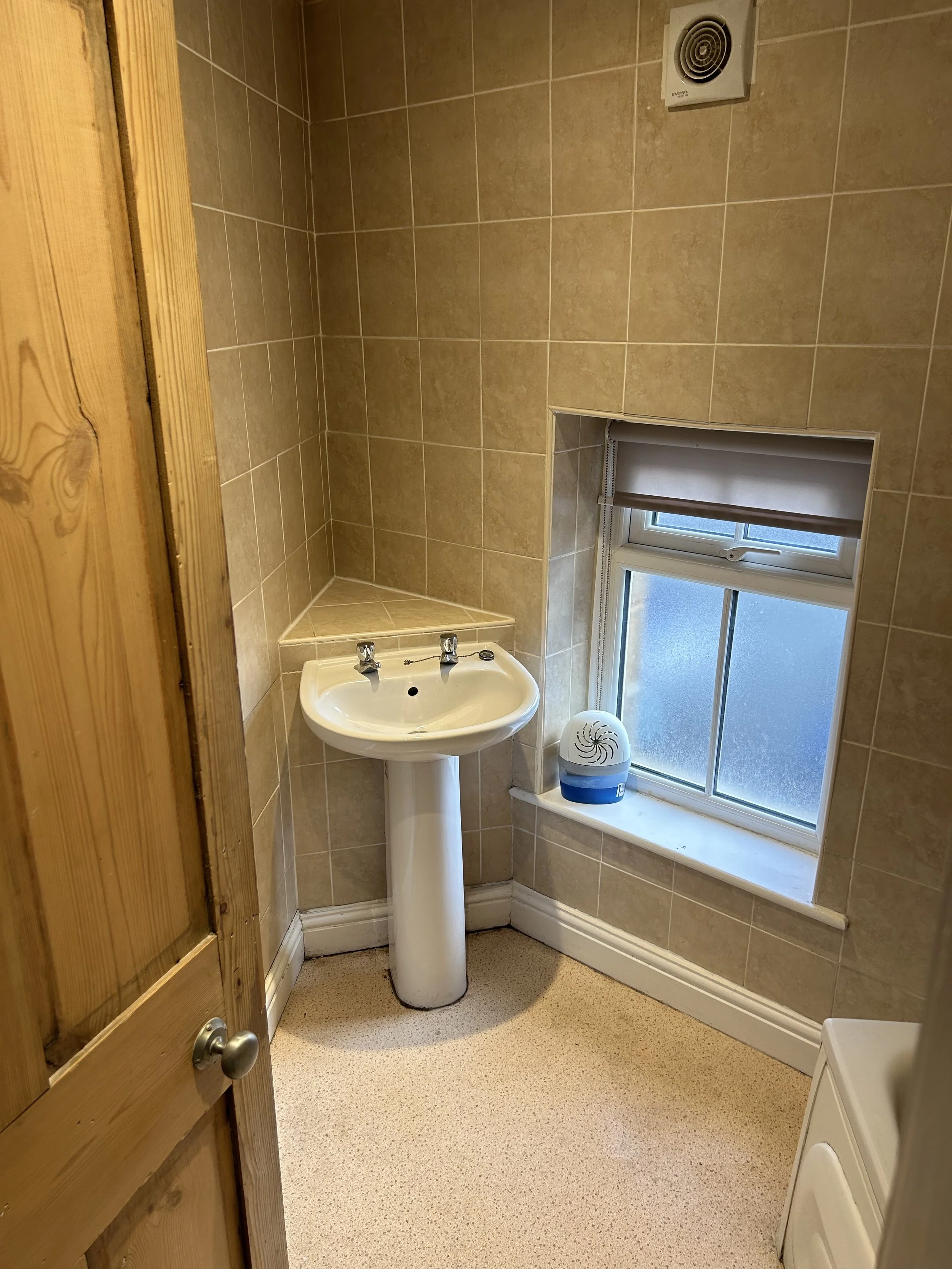 Small bathroom with beige tiled walls, a white pedestal sink, a window with a frosted glass pane, and a small space heater on the windowsill.