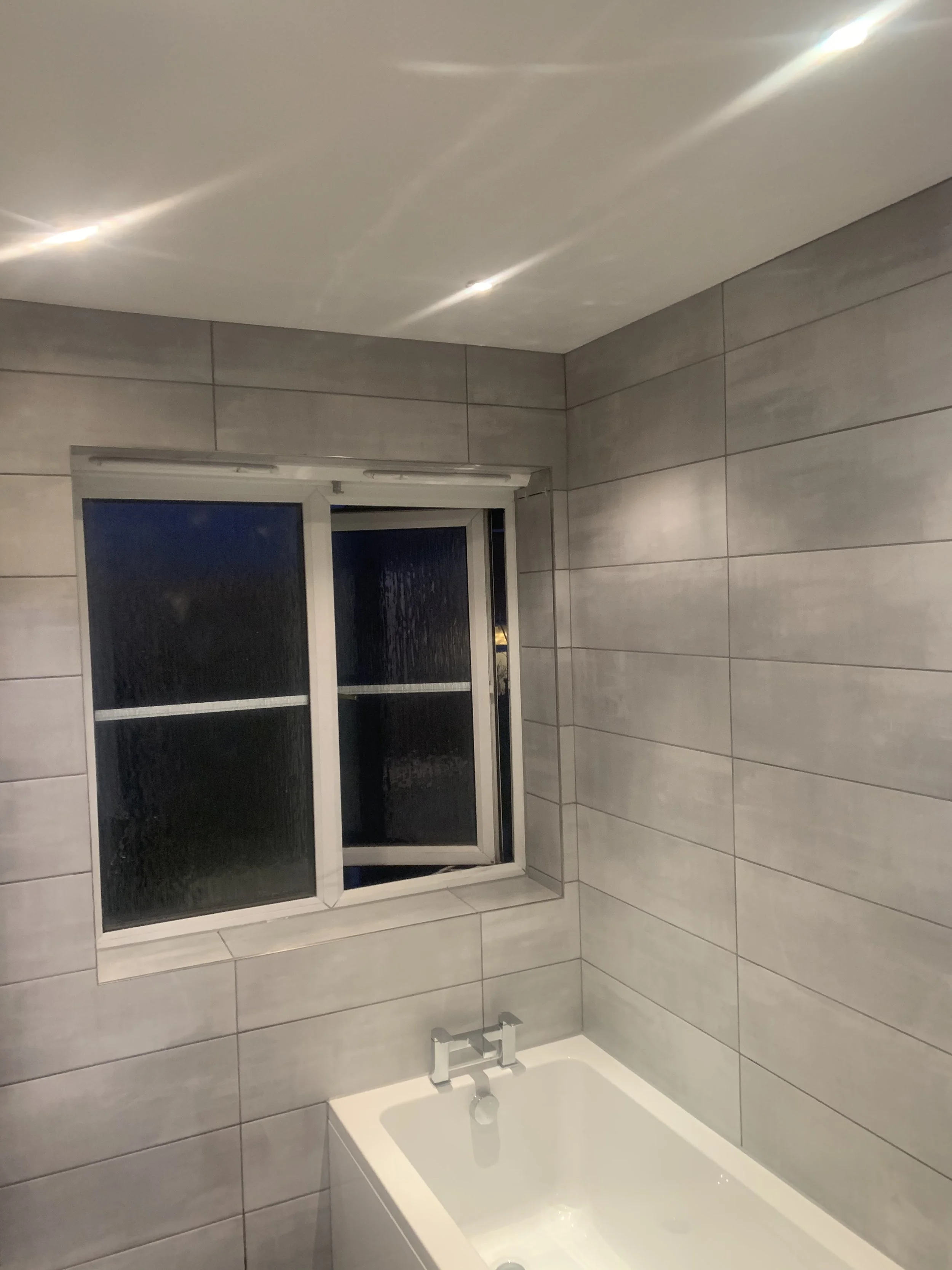 Modern bathroom with gray tiled walls, a white bathtub, a silver faucet, and a small window with frosted glass at night.