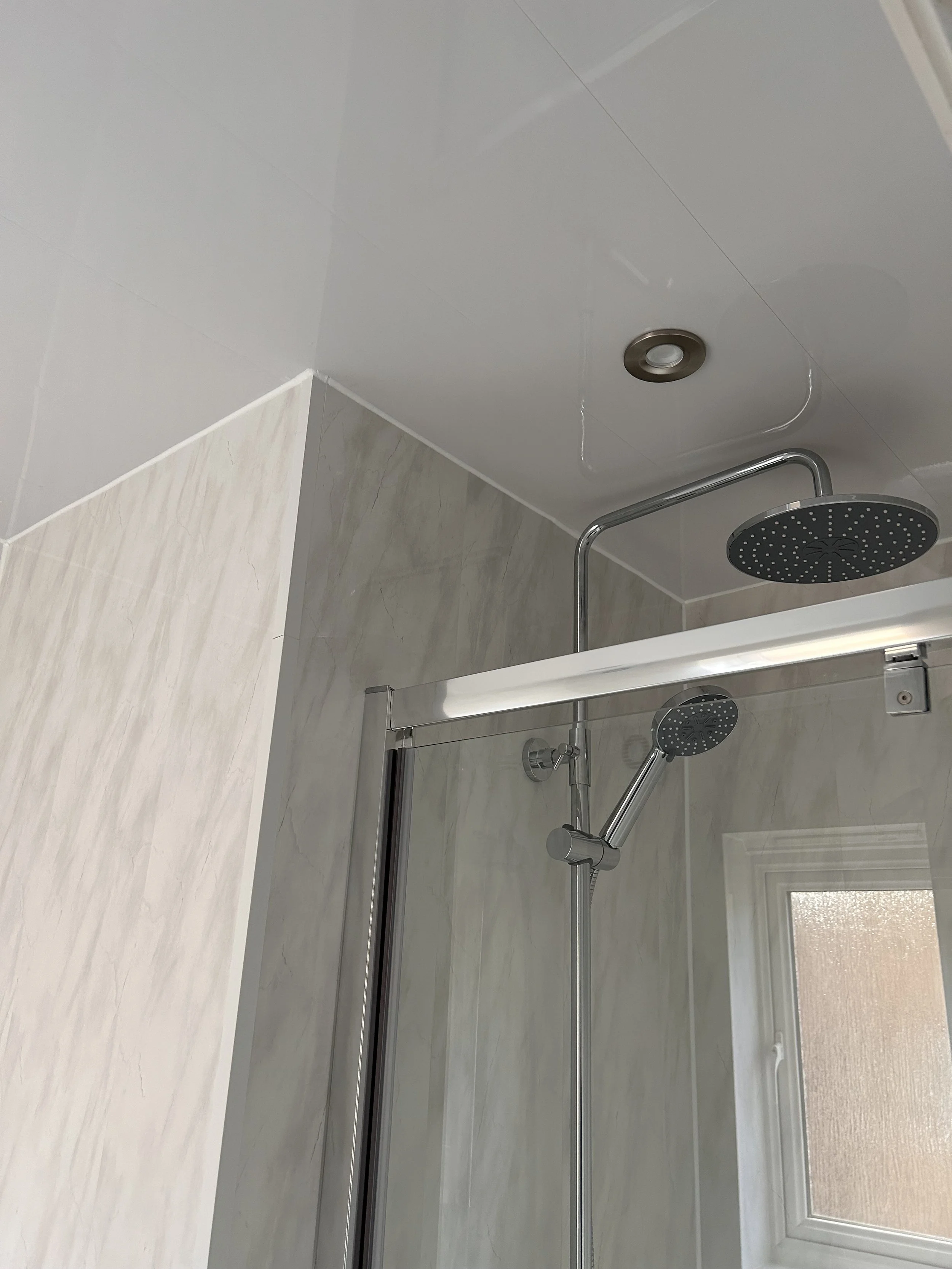 Bathroom shower area with a glass door, a round overhead showerhead, and a handheld showerhead, with a small window in the background.