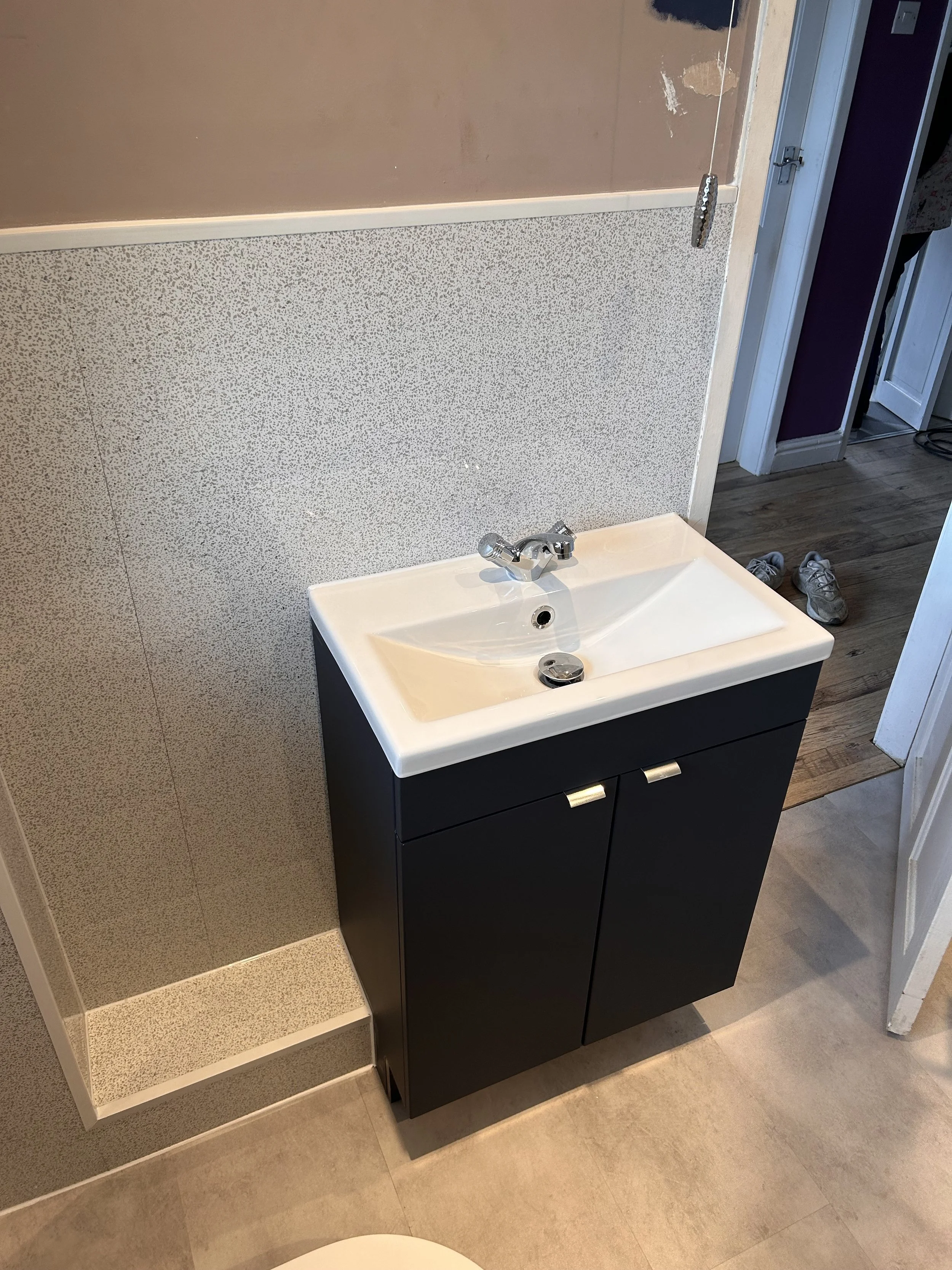 Small bathroom sink with black cabinet, white porcelain basin, and silver faucet, positioned against a speckled beige wall with a small ledge underneath, and shoes on wooden flooring nearby.