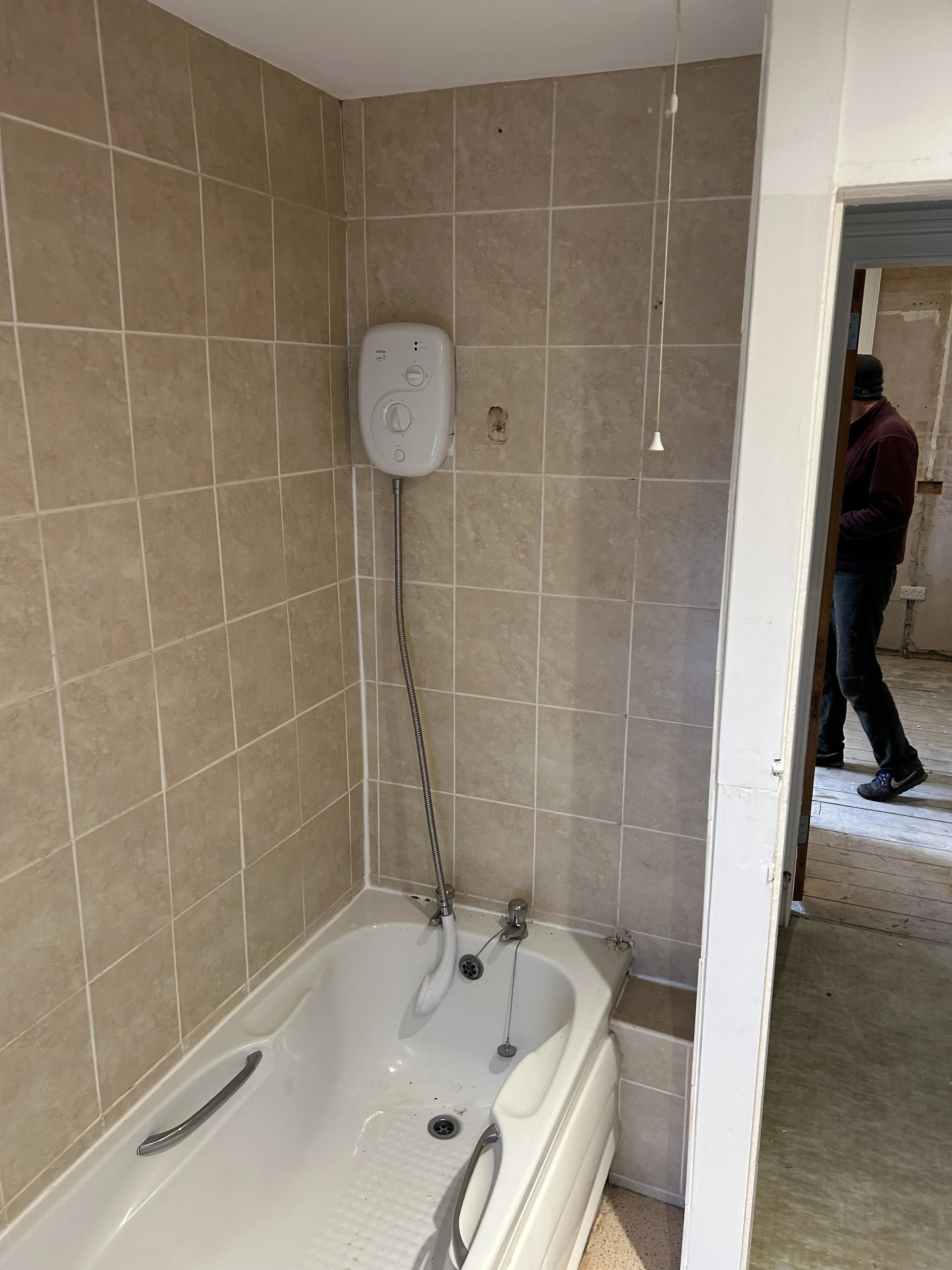 A small bathtub with handrails, a wall-mounted water heater, and beige tiled walls in a bathroom under renovation.