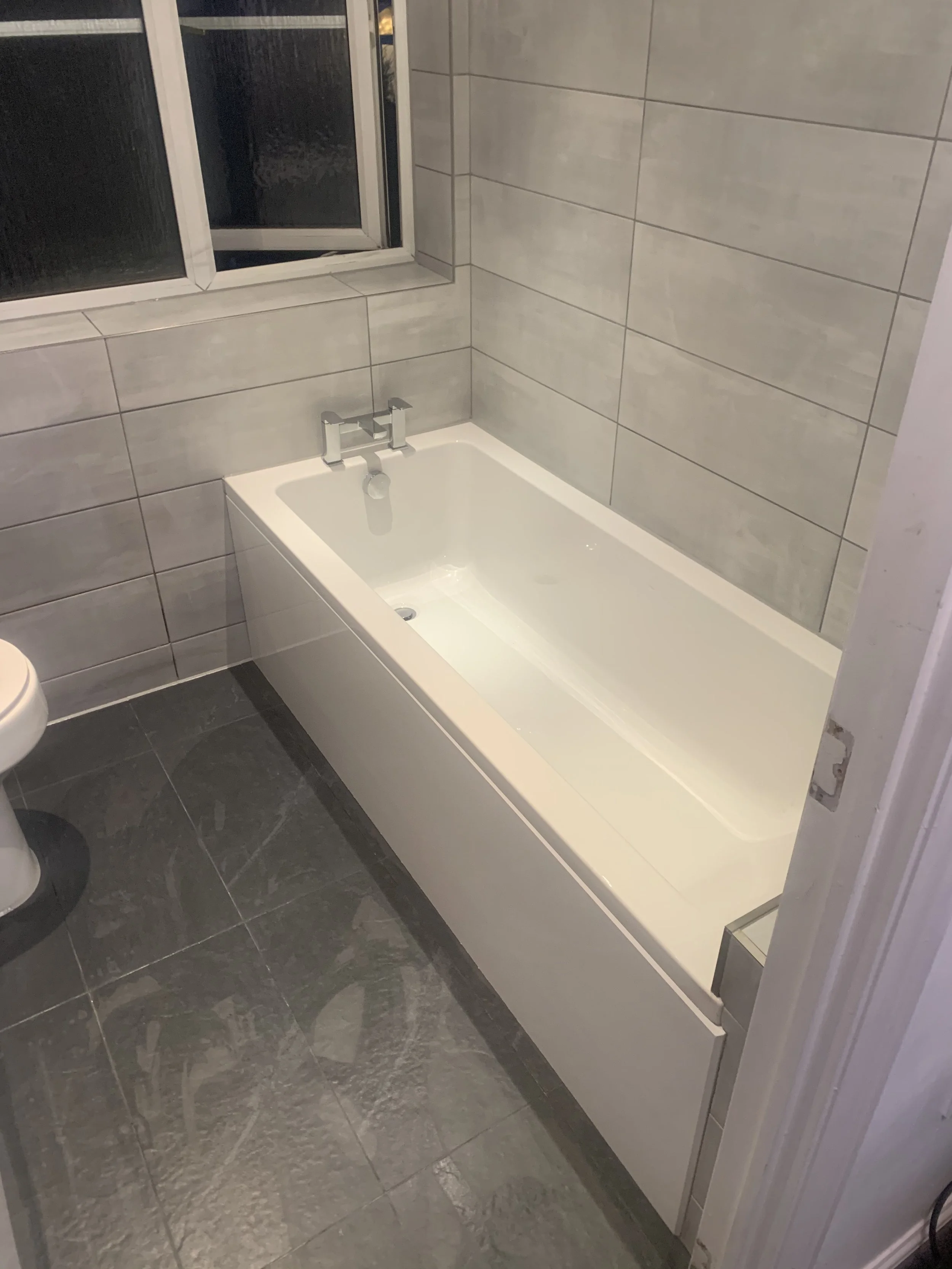 A bathroom with a white bathtub, gray tiled walls, a window, and a gray tiled floor.