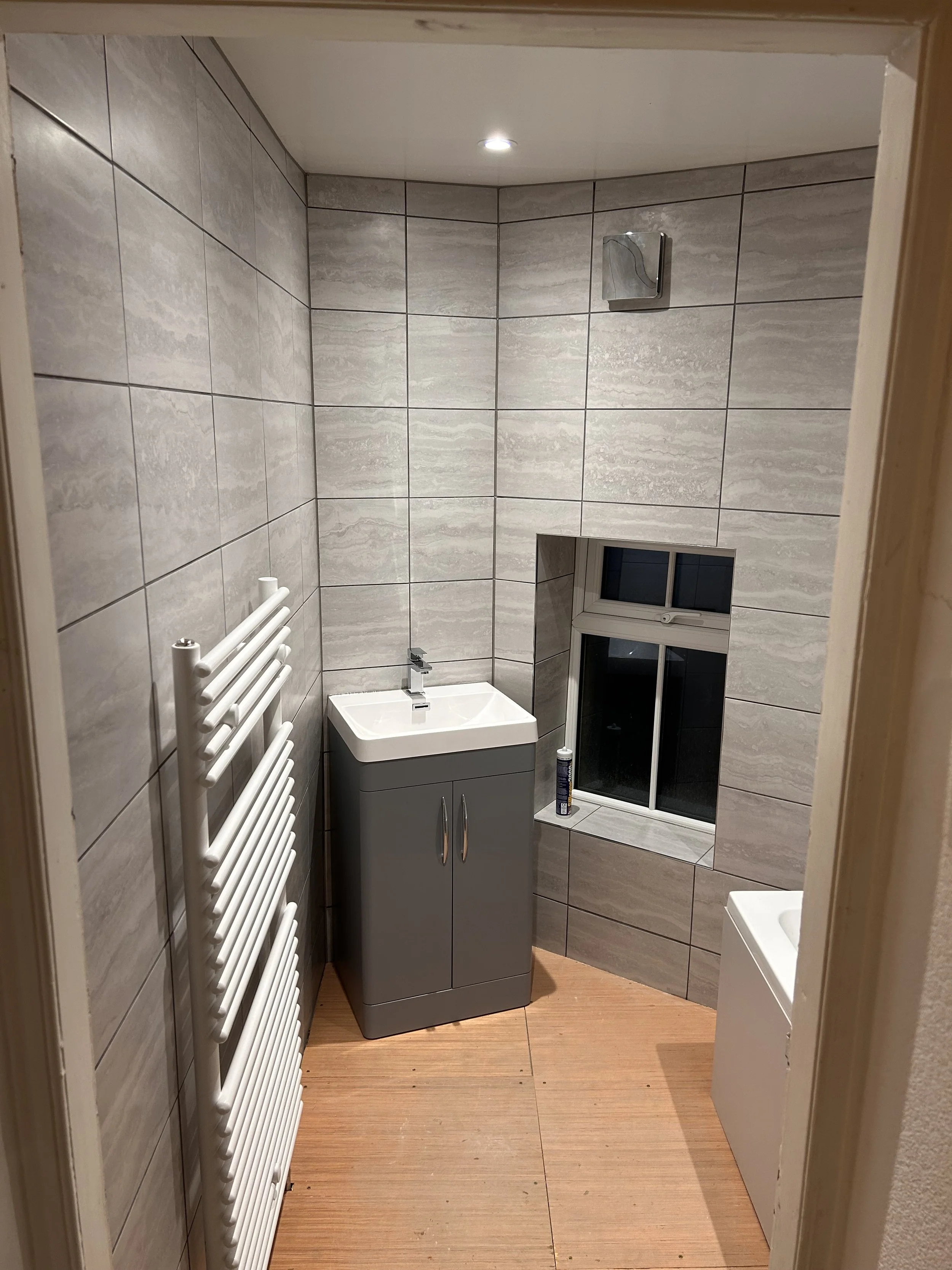 A small bathroom with light gray tiled walls, a white radiator, a gray cabinet with a sink on top, a small window with a ledge, and a white basin on the right side. There is a can placed on the window ledge and a ventilation fan on the wall near the 