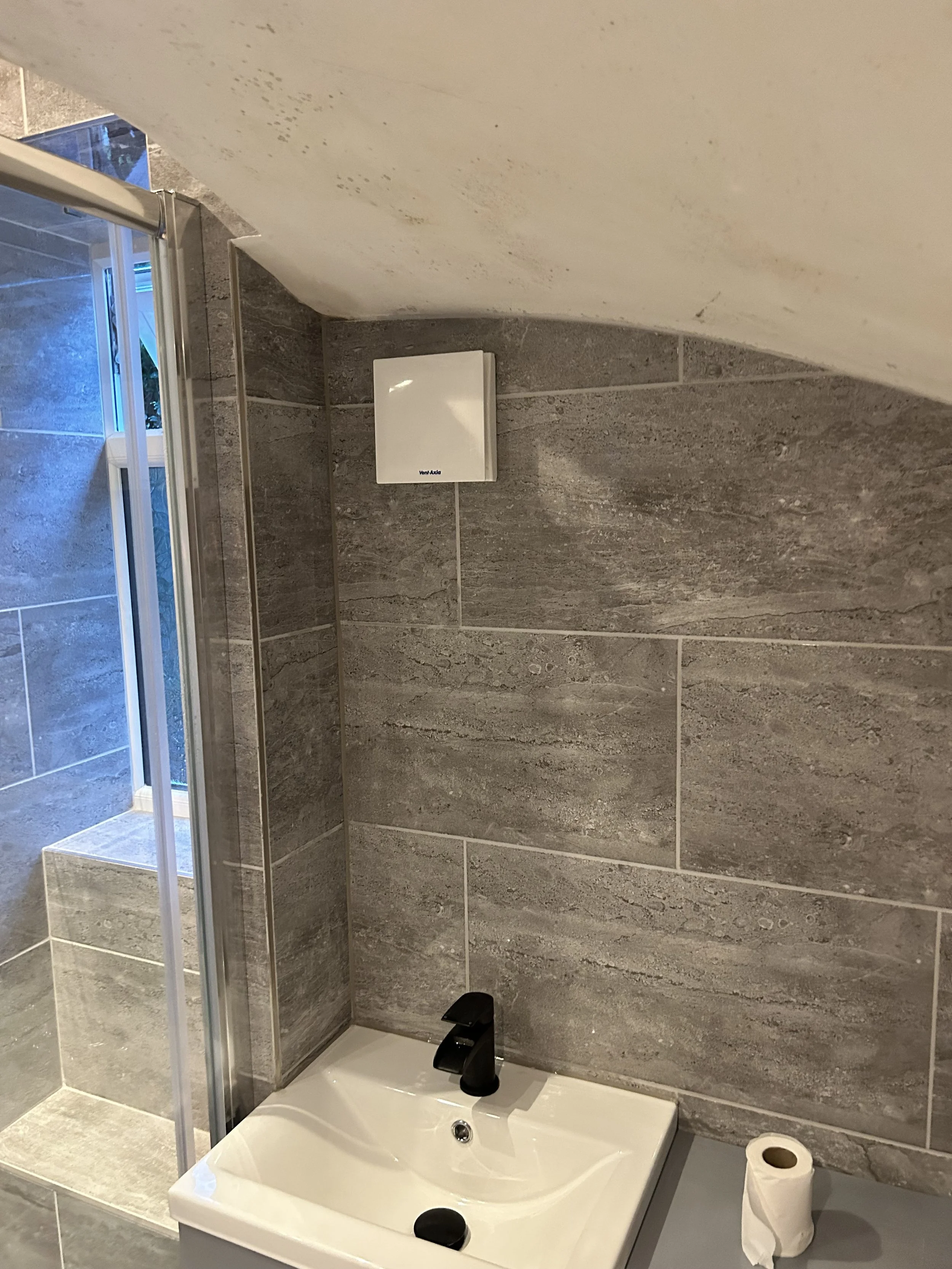 Bathroom with gray tiled wall, small white sink with black faucet, toilet paper roll on the countertop, and a shower area with glass door.