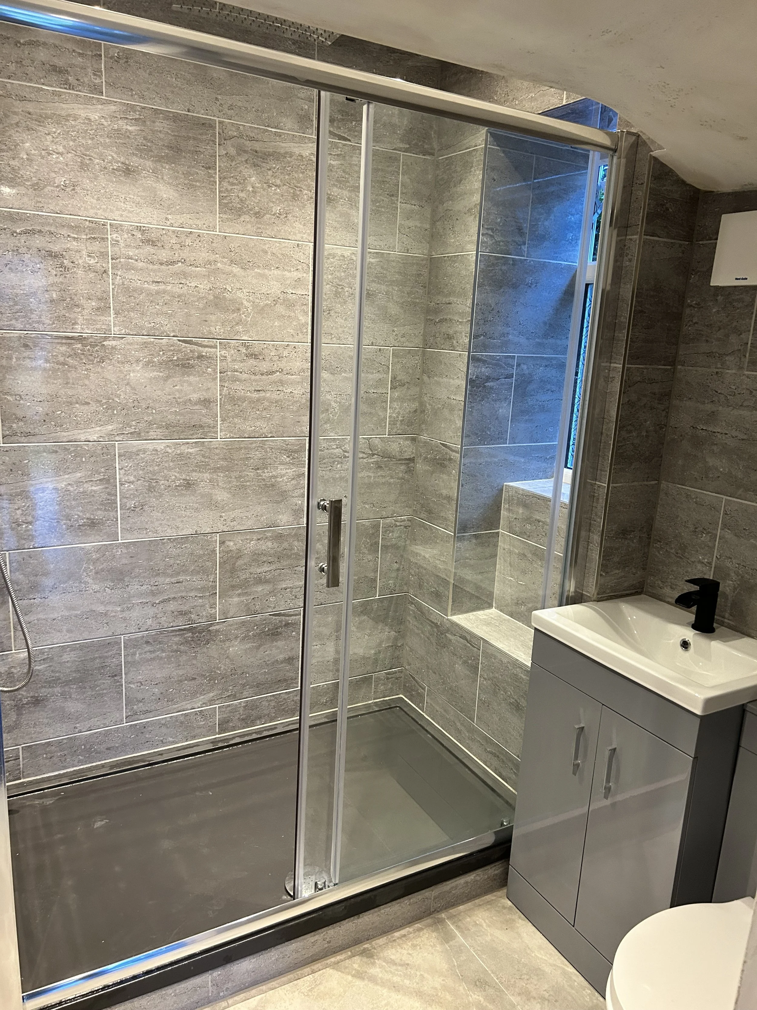 Modern bathroom with a glass-enclosed shower, gray tiles on the walls, a small window allowing natural light, a gray vanity with a white sink and black faucet, and a partial view of a toilet.