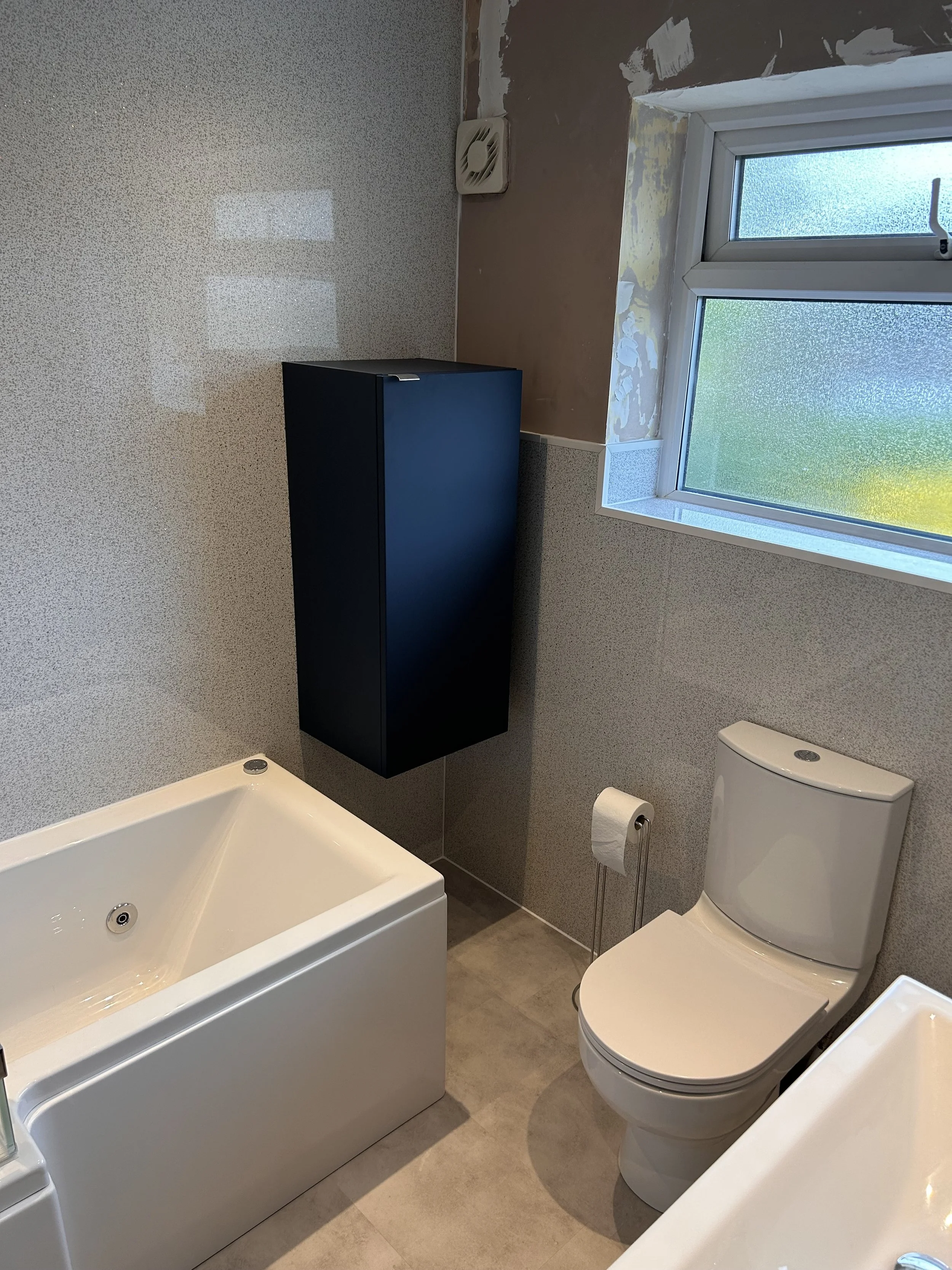 A bathroom with a bathtub, toilet, a blue wall-mounted cabinet, a window, and partially finished wall with exposed drywall.