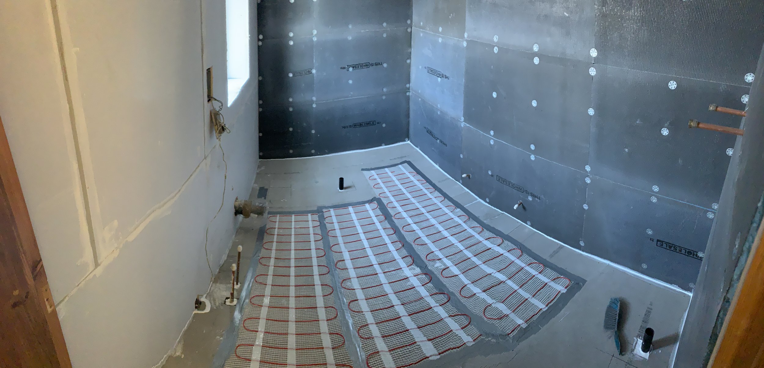 Bathroom under construction with radiant floor heating system installed, metal wall panels, exposed pipes, and construction tools.