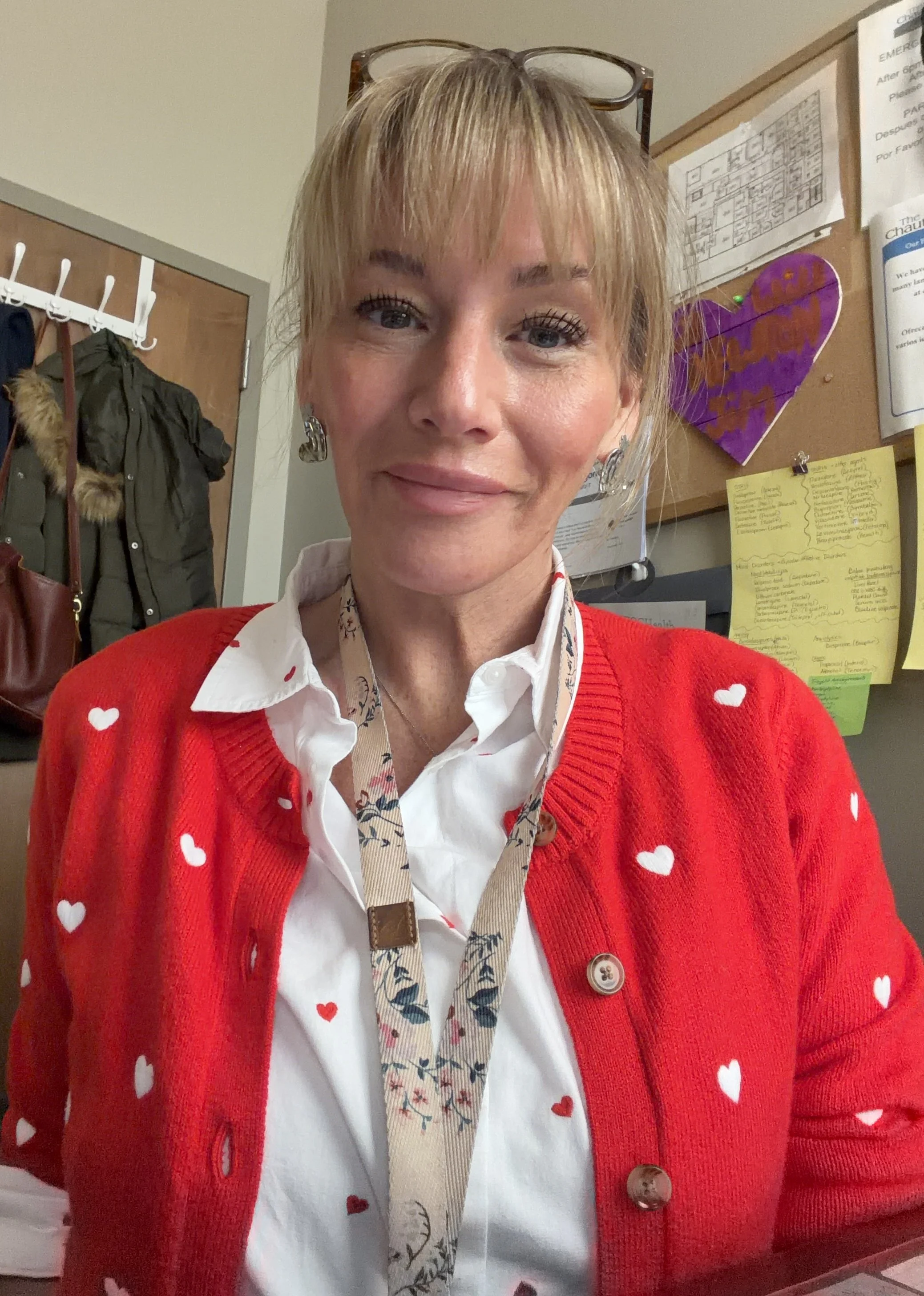 A woman with blonde hair and bangs, wearing glasses on her head, a white shirt with small red hearts, and a red cardigan with white hearts. She is in an office with a bulletin board, papers, and jackets hanging in the background.