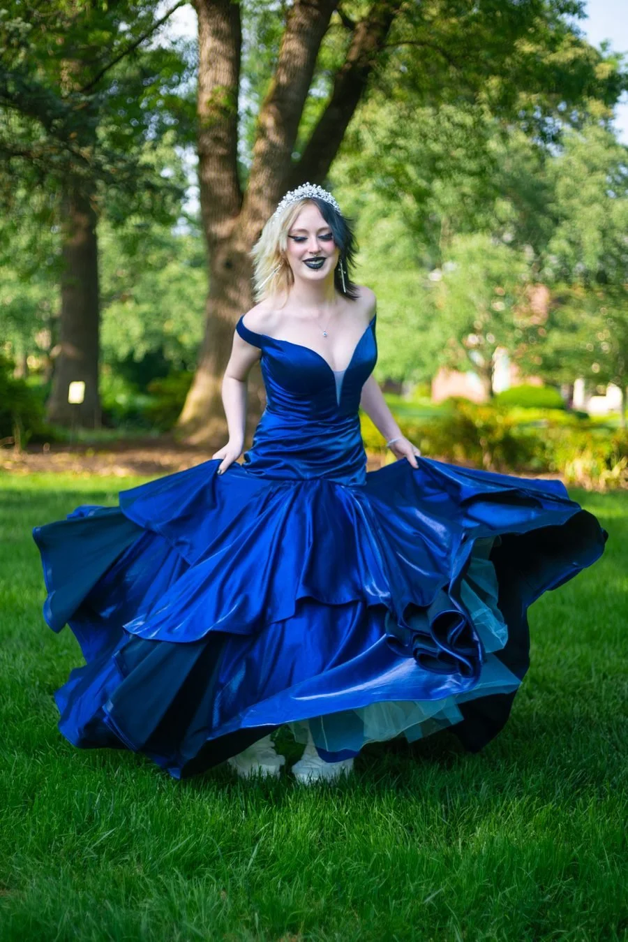 Young woman wearing a royal blue ball gown and a tiara, standing on grass in a park with trees in the background.